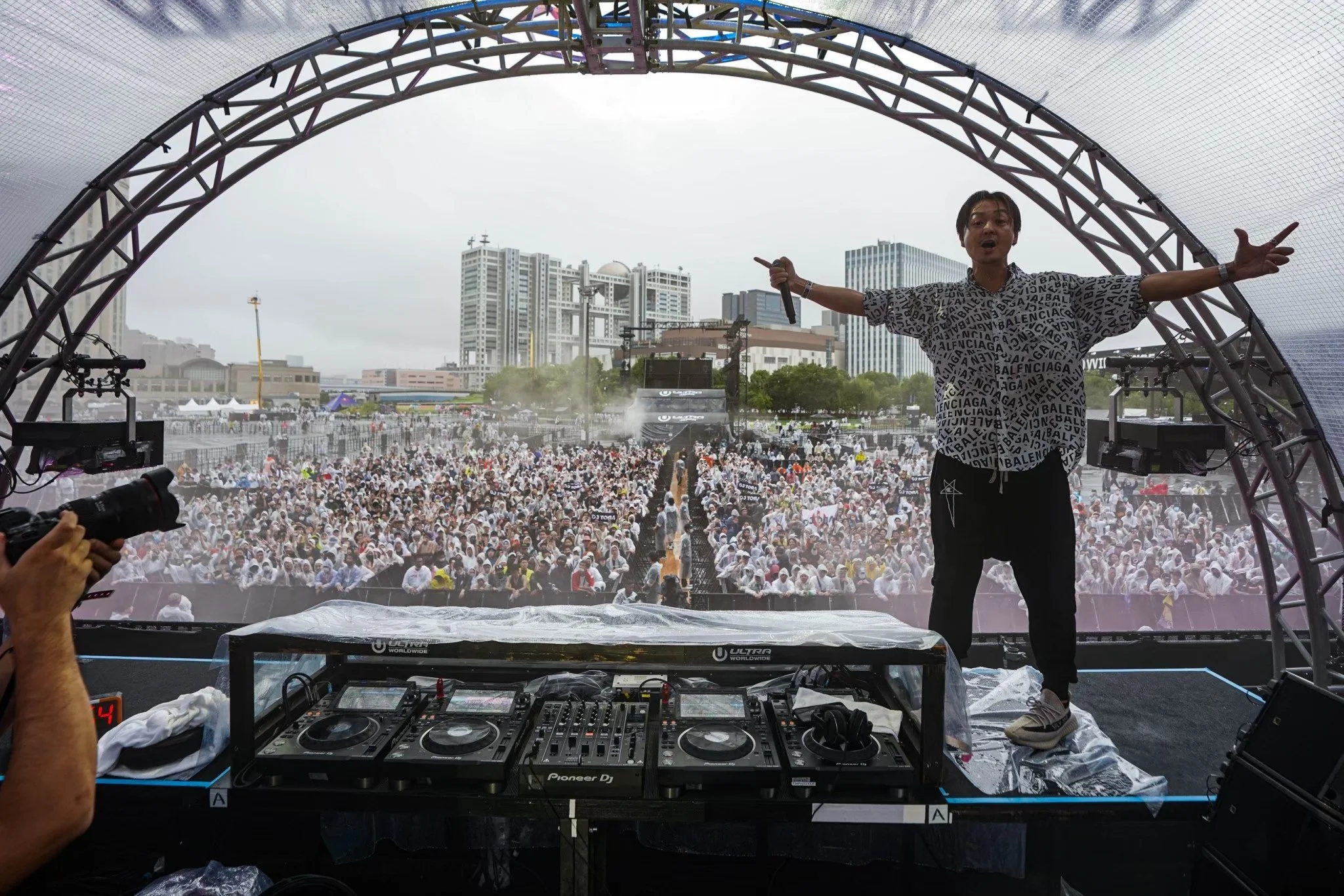 Musician standing on stage with arms outstretched, performing in front of a large crowd at an outdoor concert with city buildings in the background.