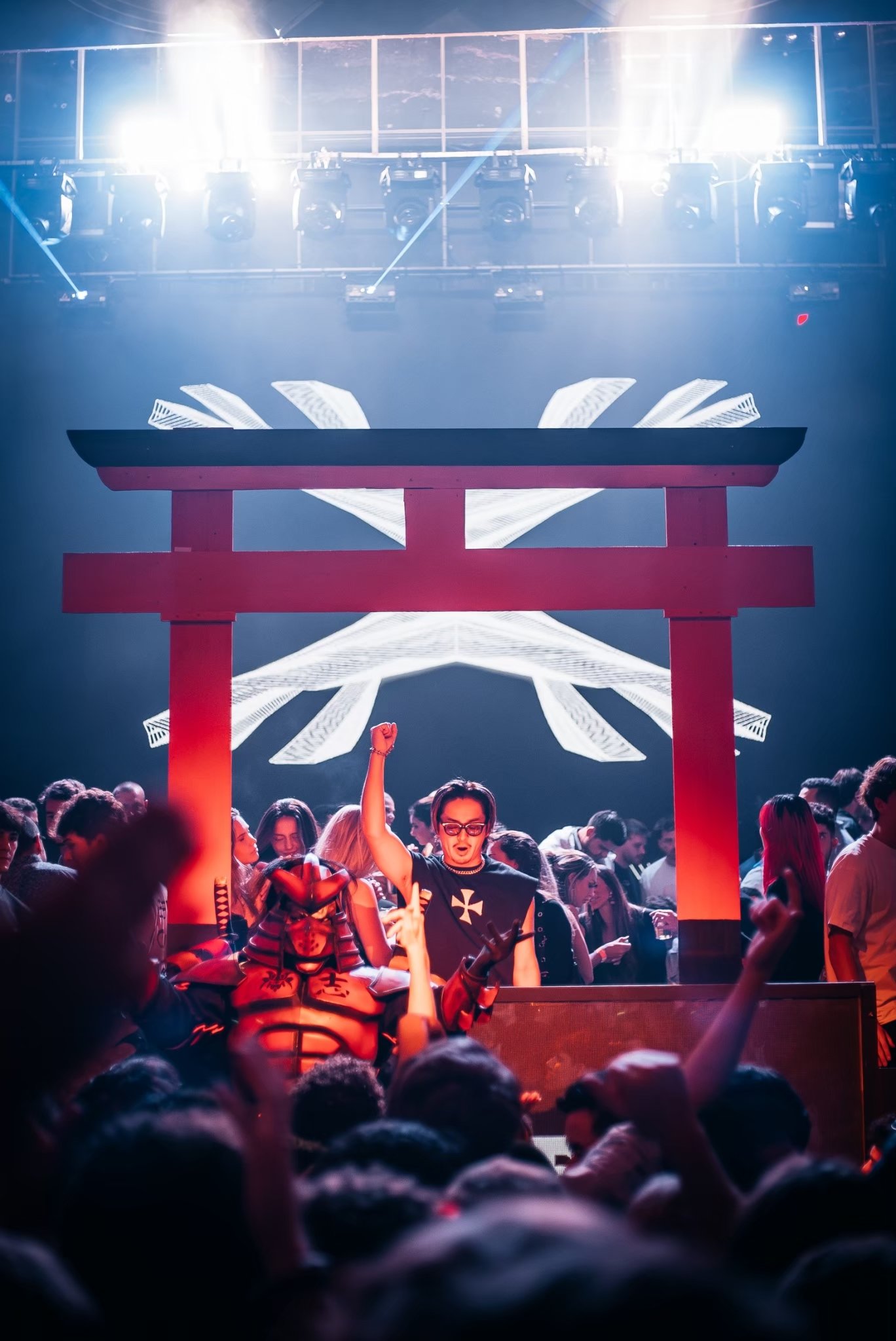 A lively nightclub scene with a crowd dancing and enjoying the music under vibrant lighting. A large red torii gate decoration is prominent in the foreground, and a bright, abstract digital display is visible behind it.