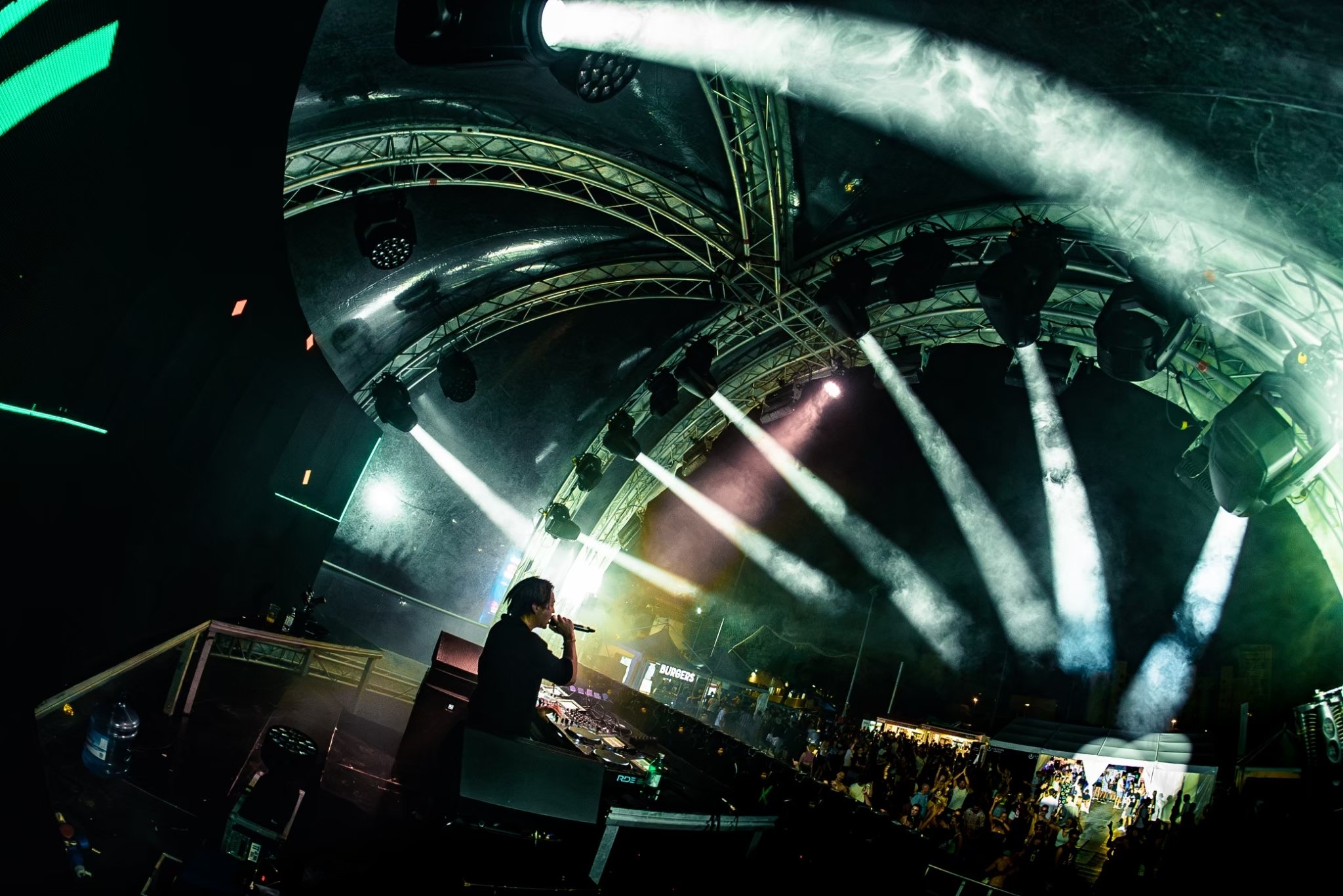 A DJ performing with a crowd at a concert under a large, arched stage with multiple stage lights and smoke effects.