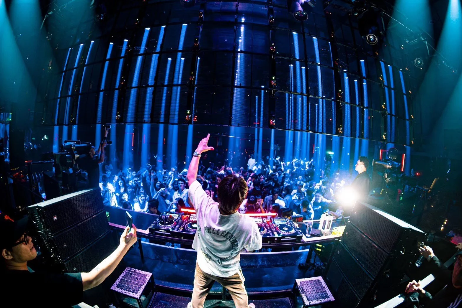 A DJ performs at a nightclub with colorful lighting and a packed dance floor. The DJ is seen from behind, wearing a white T-shirt and raising one hand. The crowd is dancing and enjoying the music, with some people taking photos or videos.