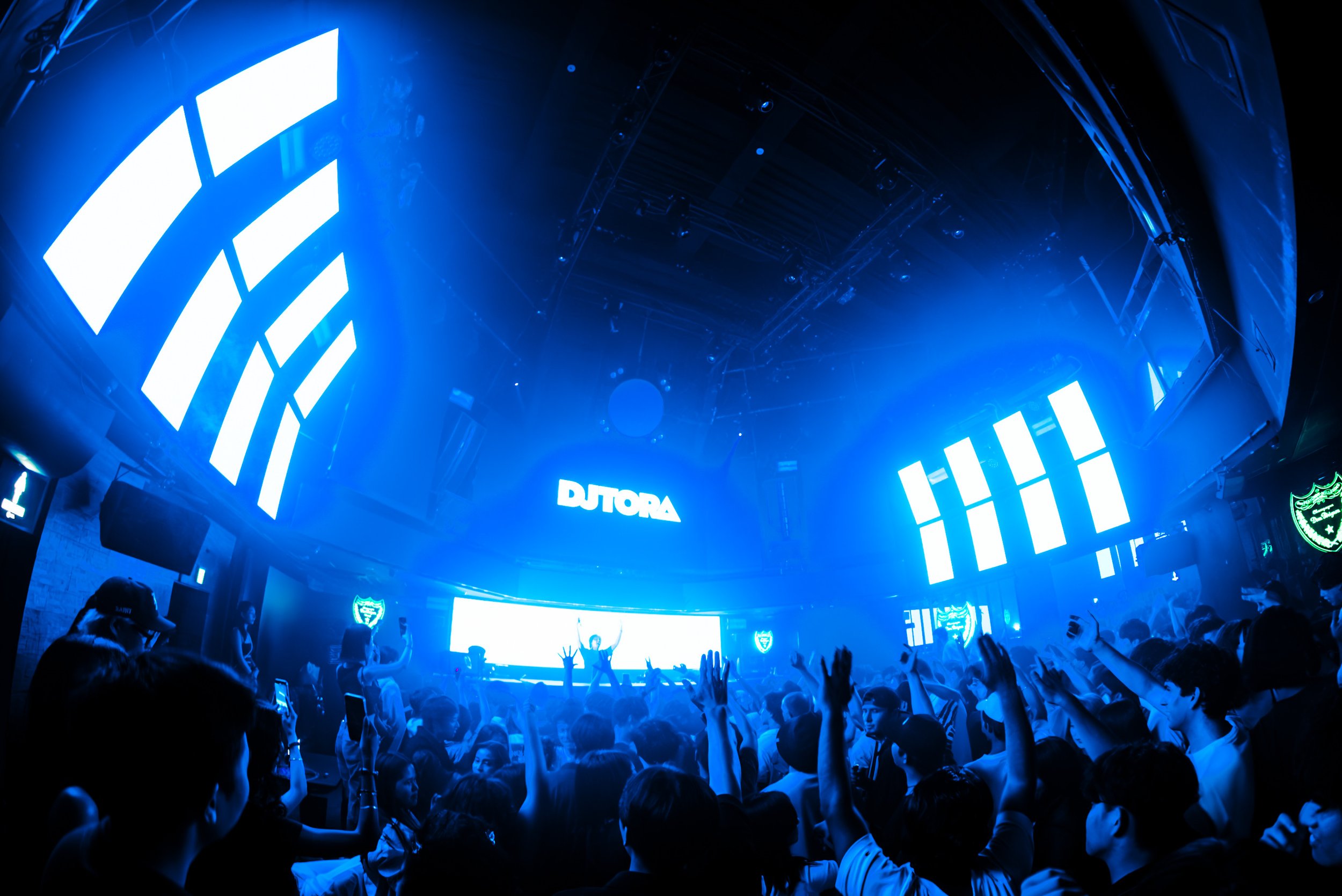 Crowd of people at a concert or event with blue lighting, raised hands, and large screens displaying the name 'DJ TORA' on stage.