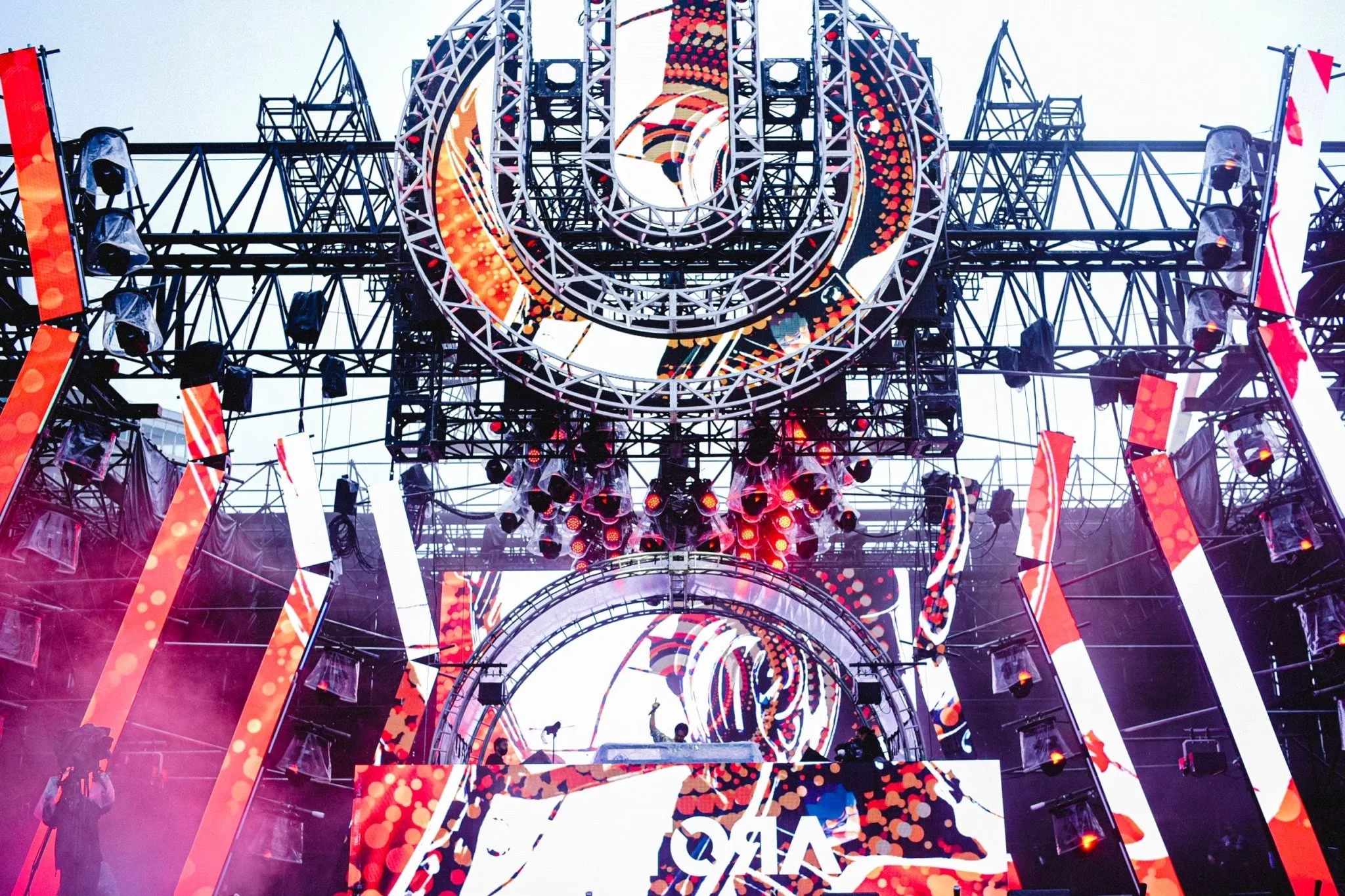 A large outdoor stage with bright LED screens and lighting equipment set up for a music event or festival, with pink and red lights creating a vibrant atmosphere.