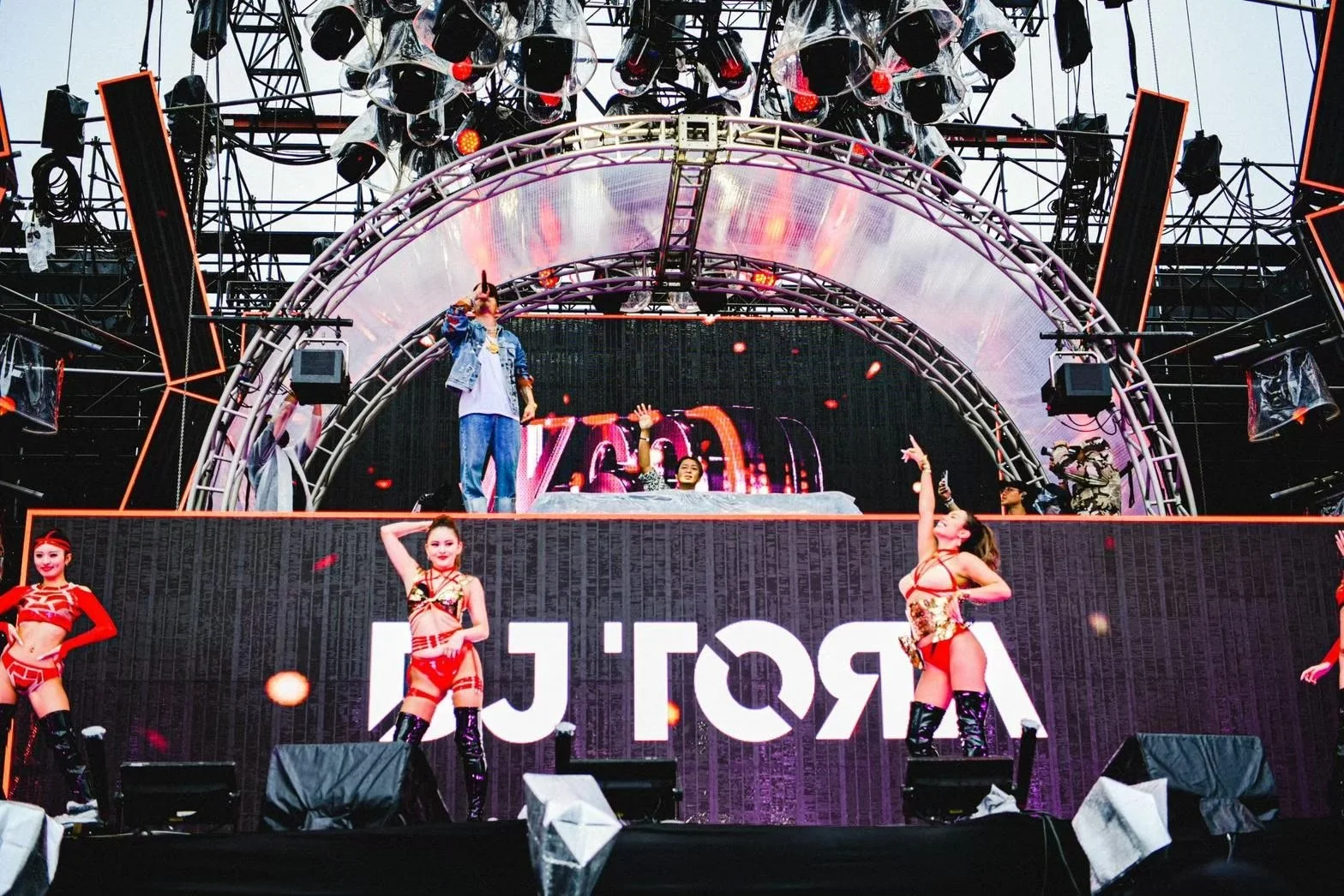 Performers on a large stage with digital screens and colorful lighting during a live concert. Four women in red and black dance costumes are in the front, while a DJ and some backup dancers are on the elevated platform behind them.