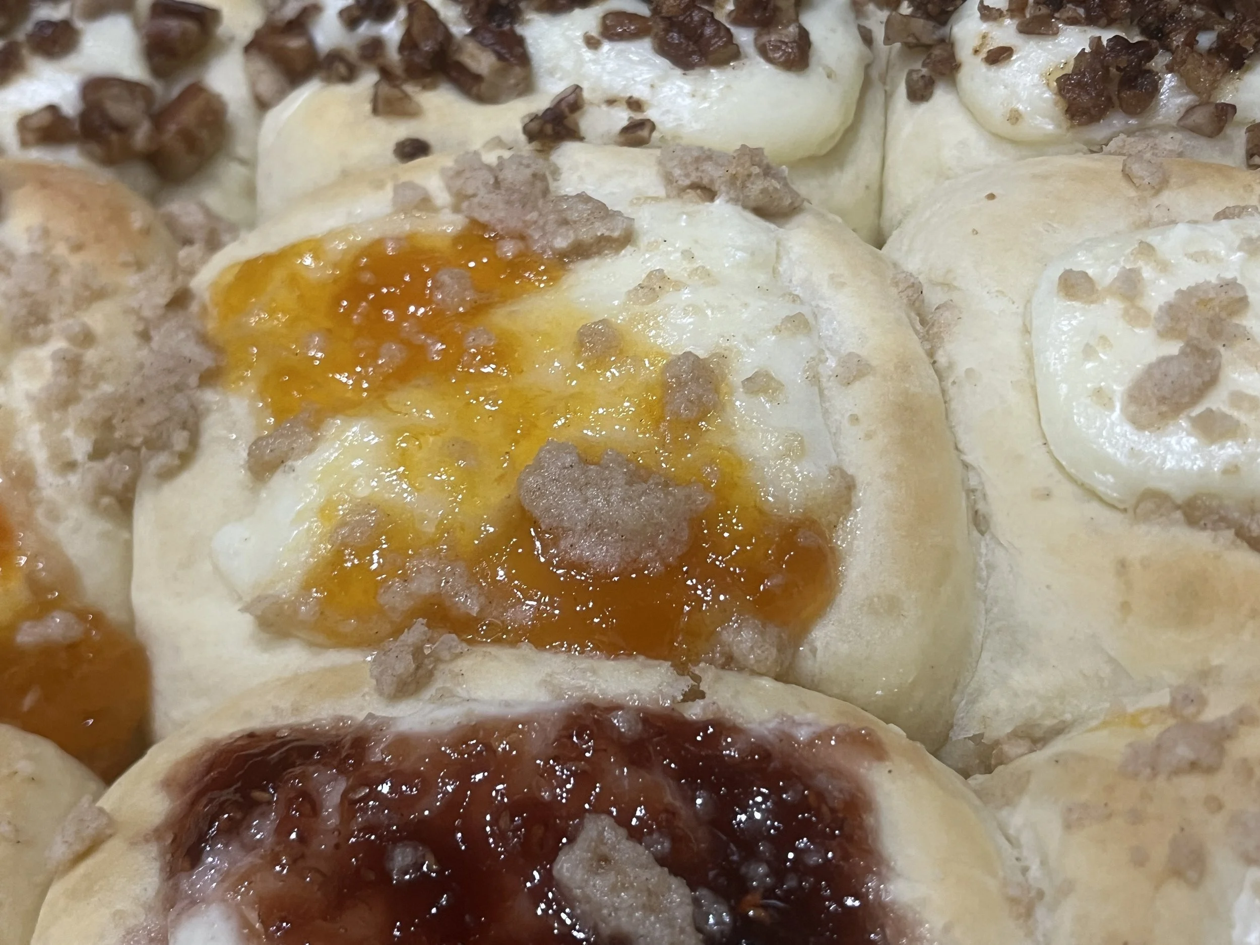 Close-up of various baked bread rolls with toppings of cheese, fruit preserves, and crumbled topping.