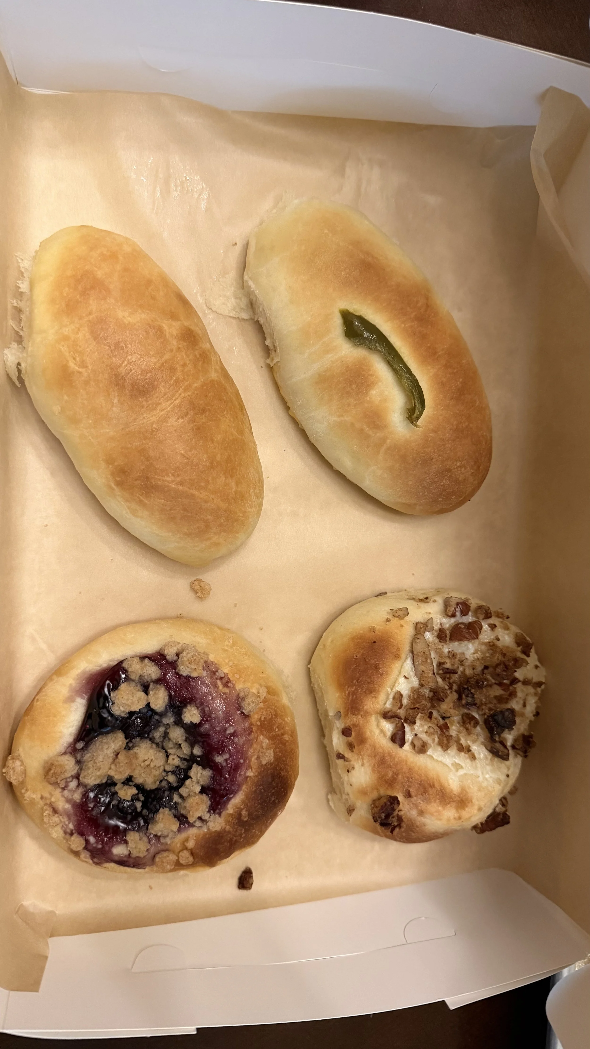 Four assorted baked pastries / kolaches in a cardboard box lined with parchment paper, including two rolls, a berry-filled pastry with crumb topping, and a pastry with nuts and dried fruit.