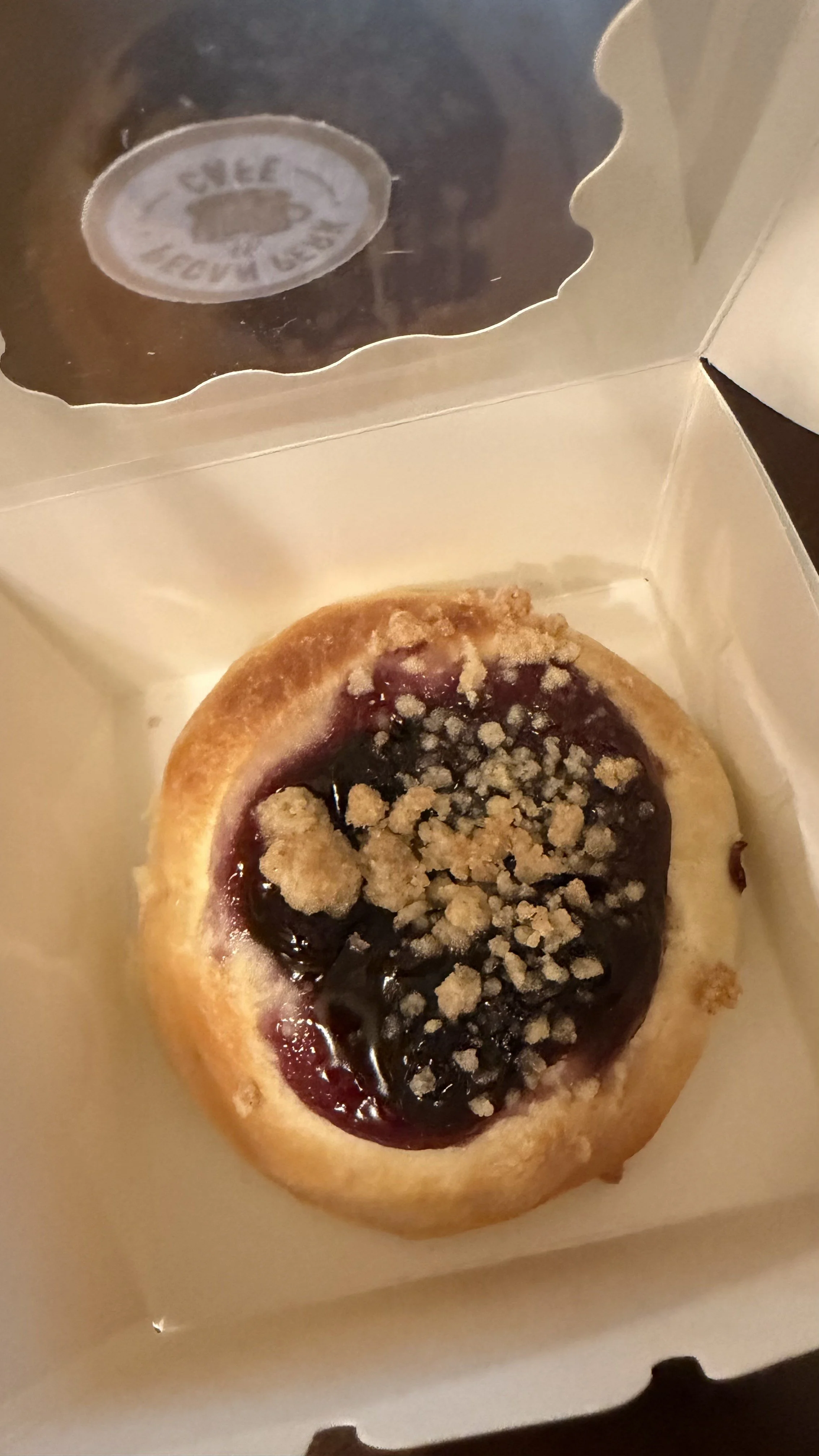 A round pastry / kolache topped with dark fruit jam and crumbled streusel, in a white box with a clear plastic window.