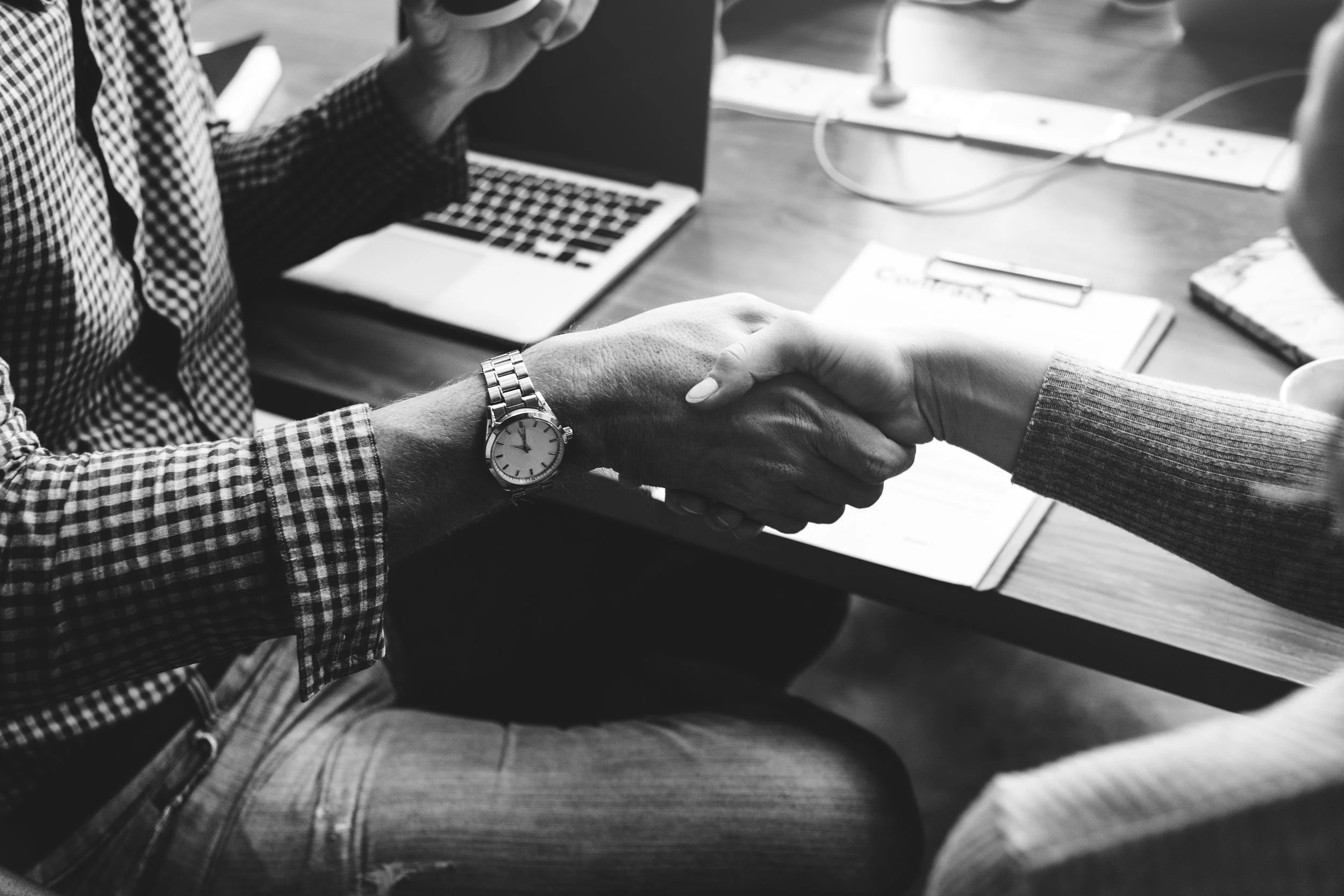 Two people shaking hands over a desk, with one wearing a watch and another in a sweater, in a business or office setting.
