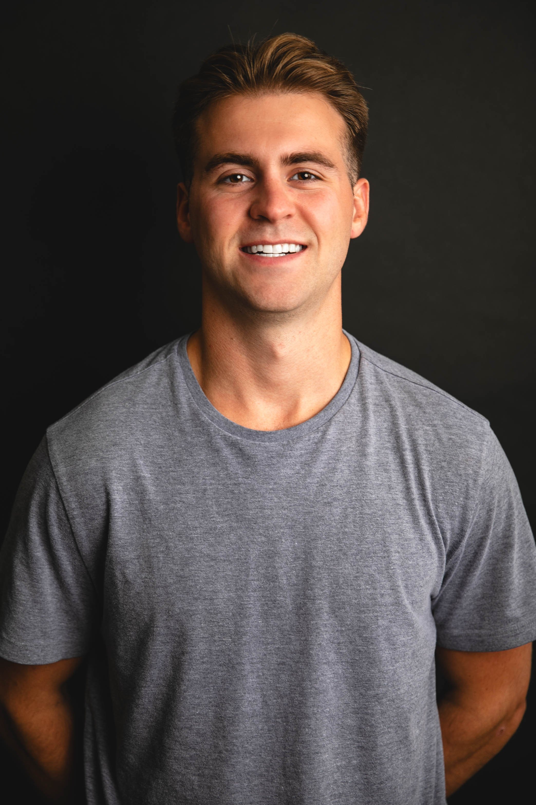 A smiling young man with short brown hair, wearing a gray t-shirt, stands in front of a black background.