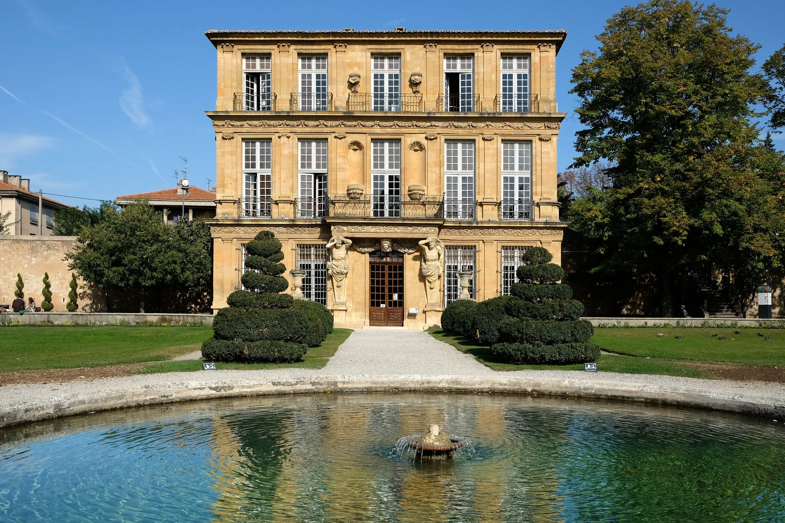 A historic four-story building in France,  surrounded by manicured trees, a gravel pathway, a fountain in the foreground, and a bright blue sky.