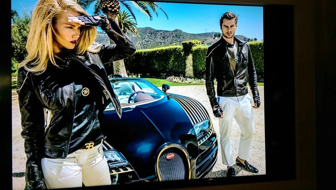 Two models in black leather jackets and white pants posing in front of a black Bugatti Chiron car outdoors on a sunny day.