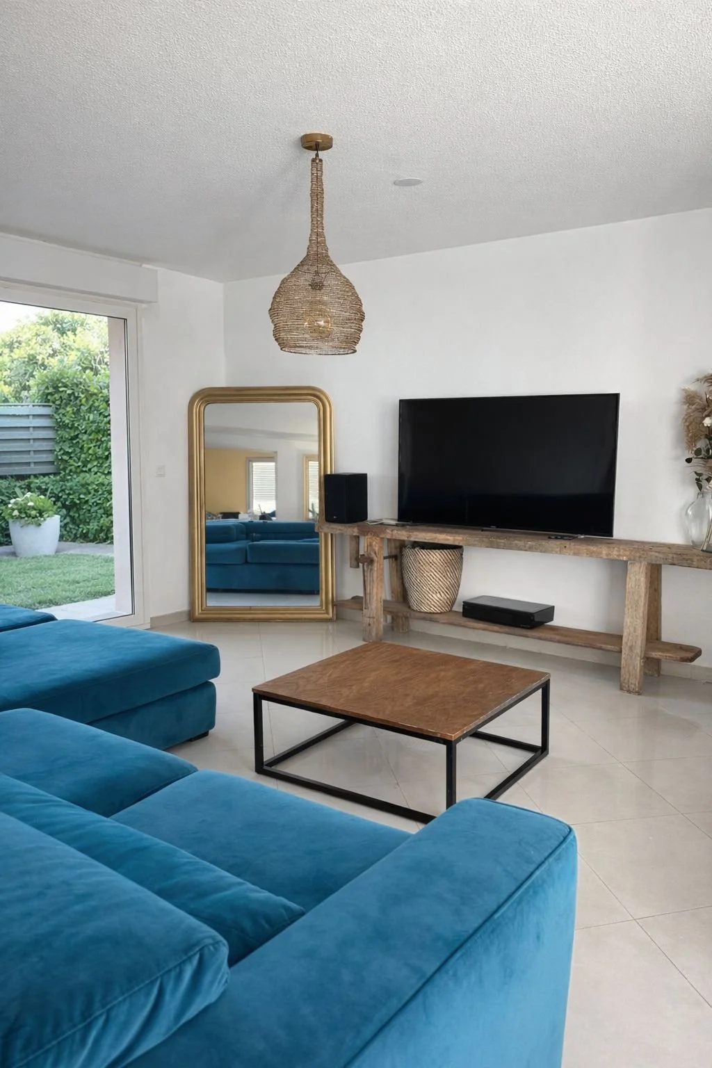 Salon moderne avec canapé bleu, télévision à écran plat sur une console en bois, miroir ancien doré, lampe suspendue en rotin, et accès au jardin extérieur
