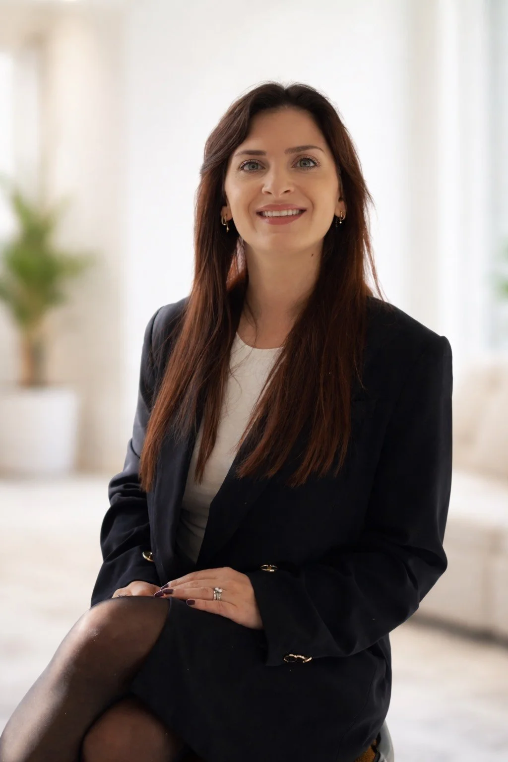 Une femme avec de long cheveux bruns, portant un blazer noir et une jupe noire, assise dans un environnement lumineux, souriant à la caméra.