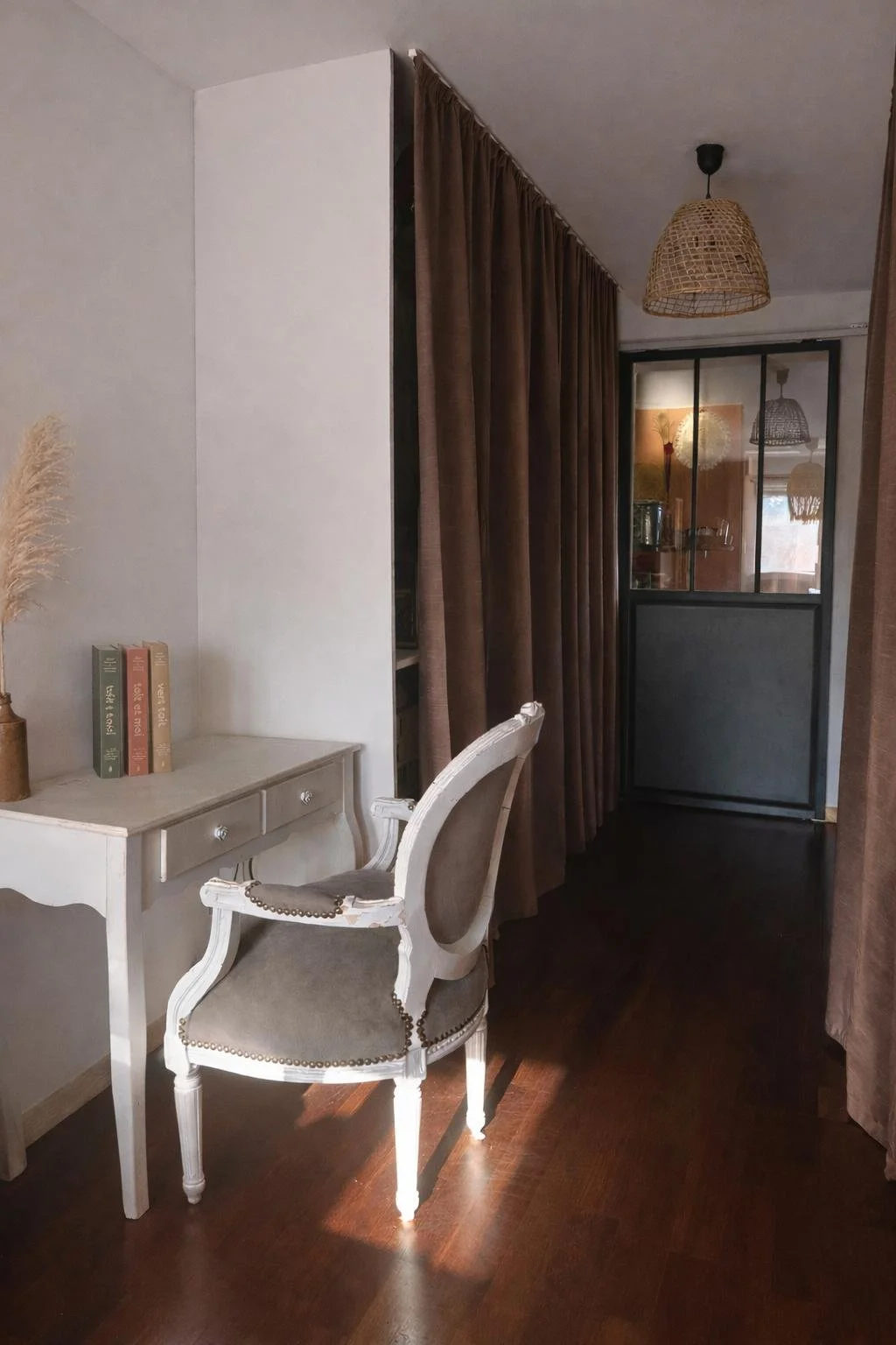 Intérieur d'une pièce avec un bureau blanc, une chaise ancienne, des livres, des rideaux marron et une porte en verre.