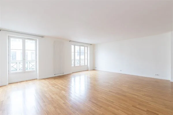 Grand salon vide avec fenêtres lumineuses, murs blancs et sol en bois.