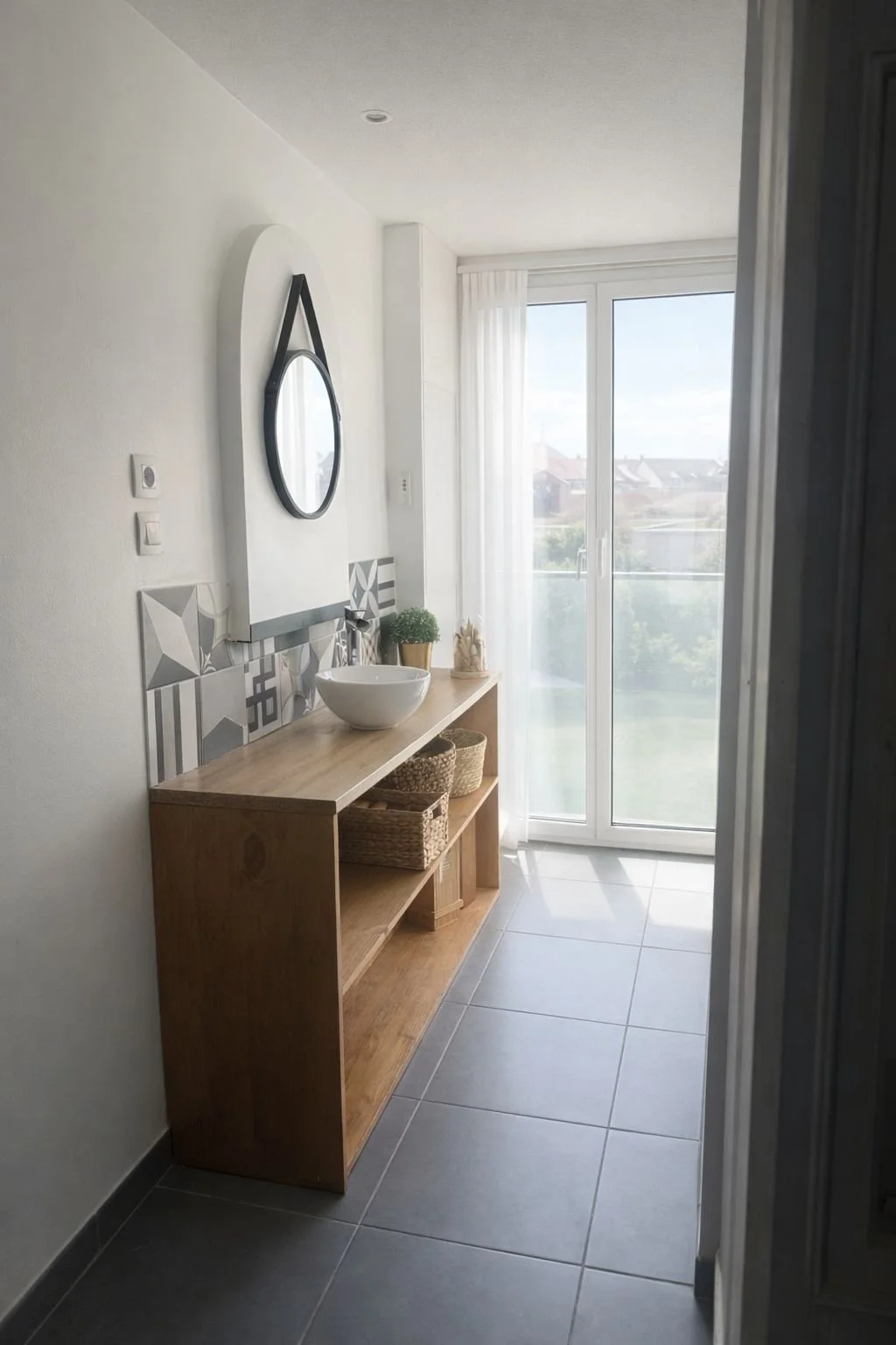 Une salle de bain lumineuse avec un meuble en bois, un grand miroir, une vasque blanche, des paniers en osier et une porte-fenêtre laissant entrer la lumière naturelle.