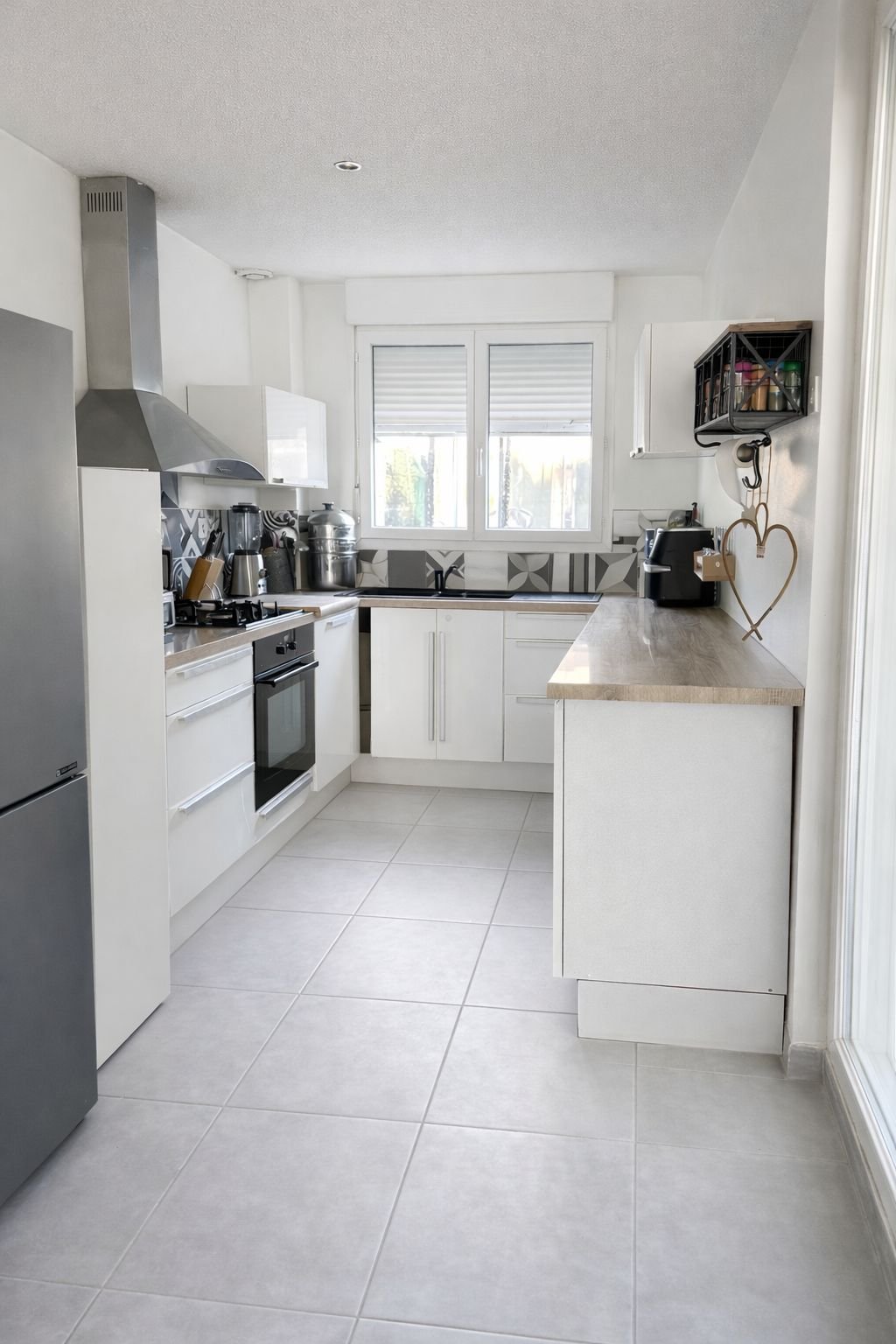 Cuisine moderne avec fenêtres, placards blancs, plan de travail en bois, appareils électroménagers en acier inoxydable, carrelage clair et décoration minimaliste.