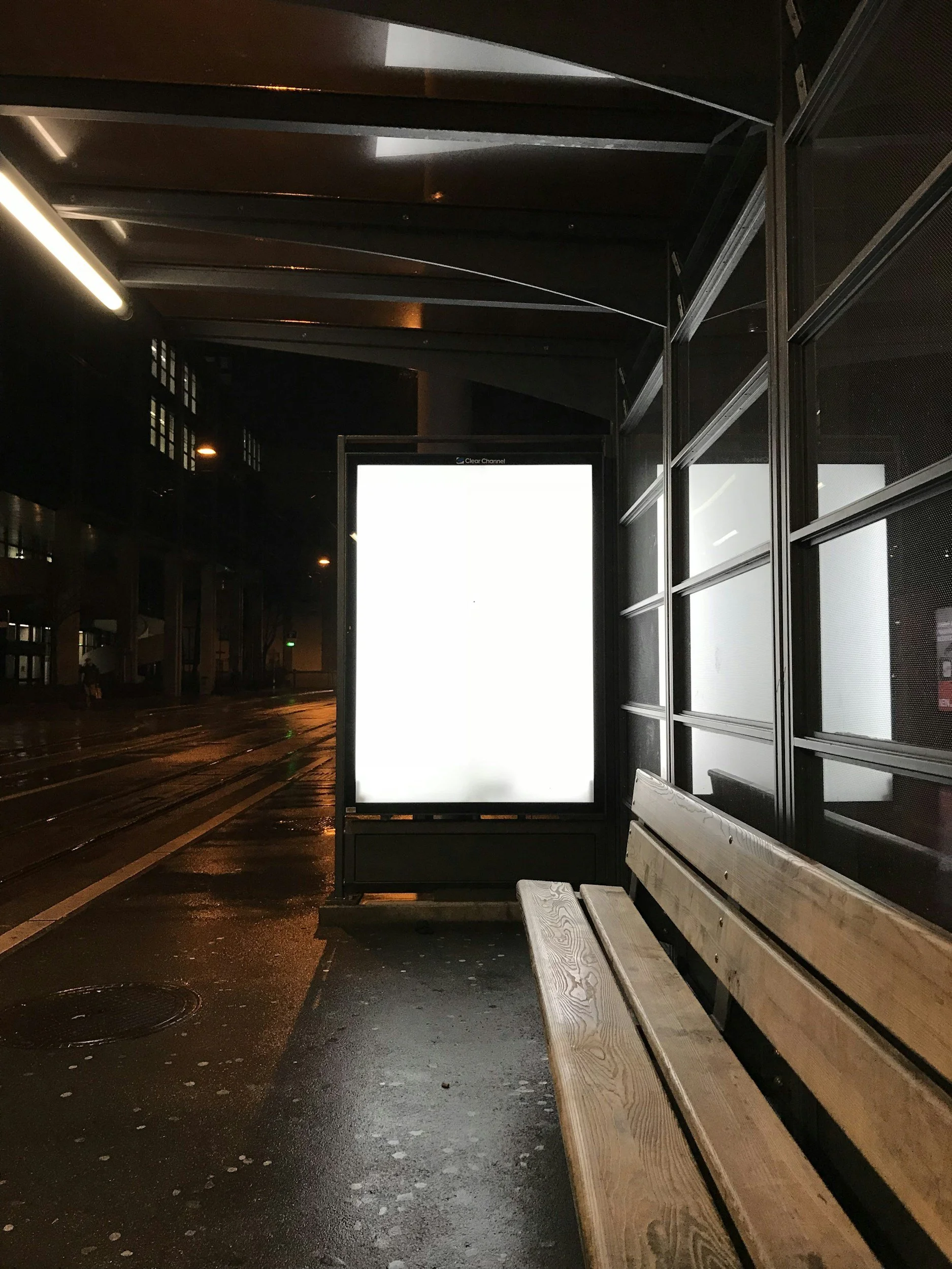 Empty bus stop bench at night with a brightly lit advertisement board and wet pavement.