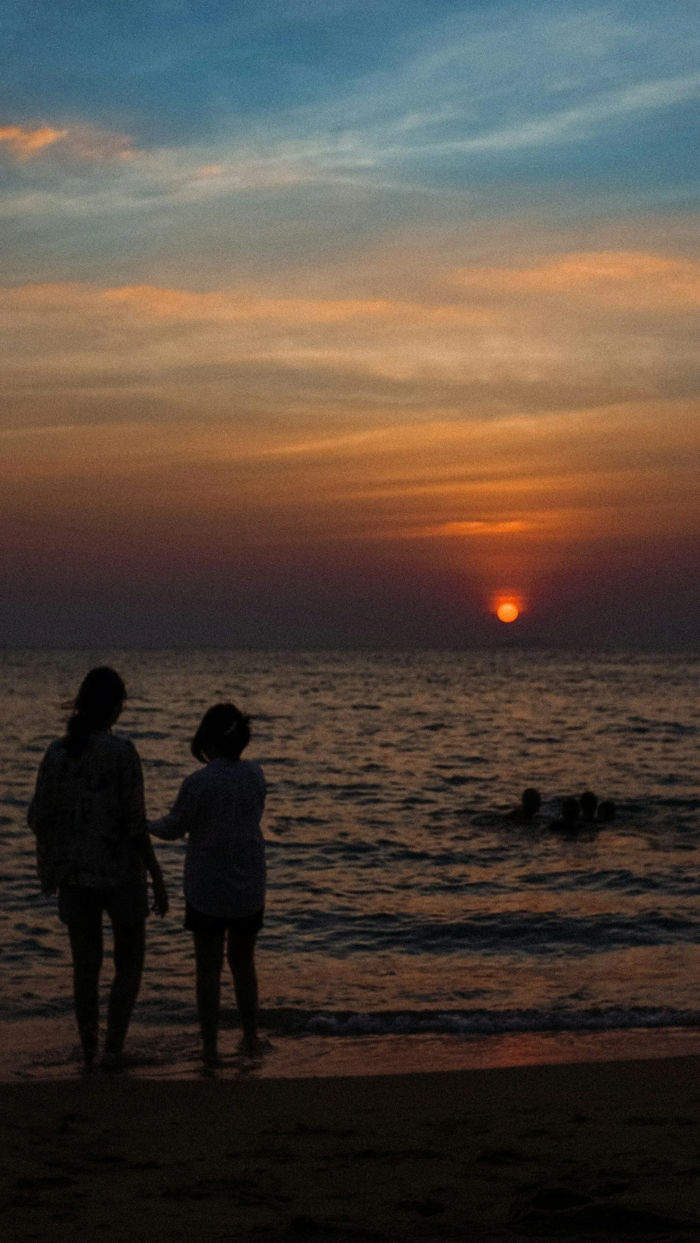 Due persone silhouette sulla spiaggia al tramonto, con il mare e il sole calante all'orizzonte, e alcuni bambini che giocano in acqua sullo sfondo.
