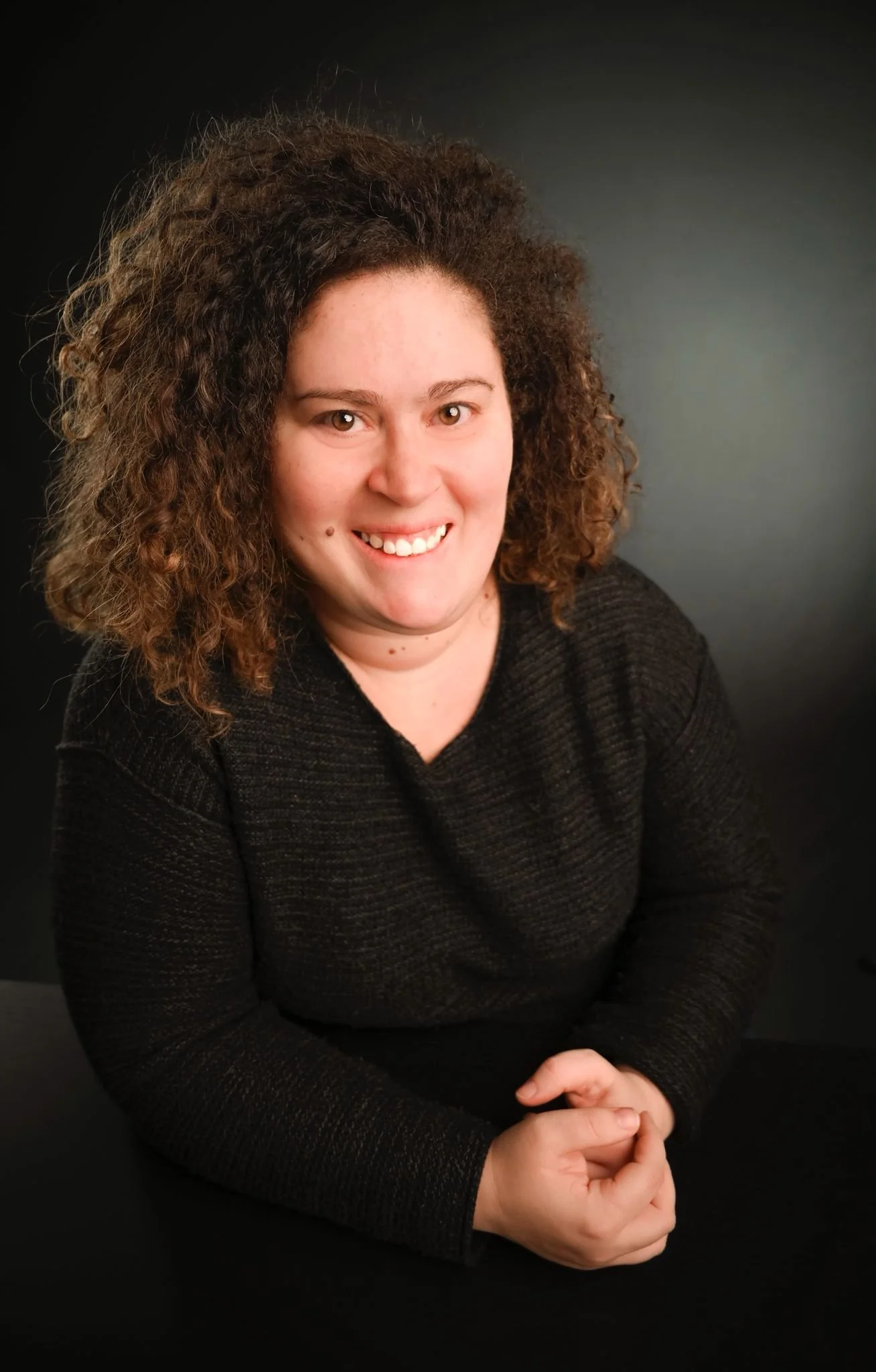 Ritratto di una donna con capelli ricci e folti, sorridente, che indossa una maglietta nera, seduta davanti a uno sfondo grigio scuro. Ingegnera, donna STEM, orientatrice ASNOR in formazione, determinata a sostenere altre donne STEM nella carriera.