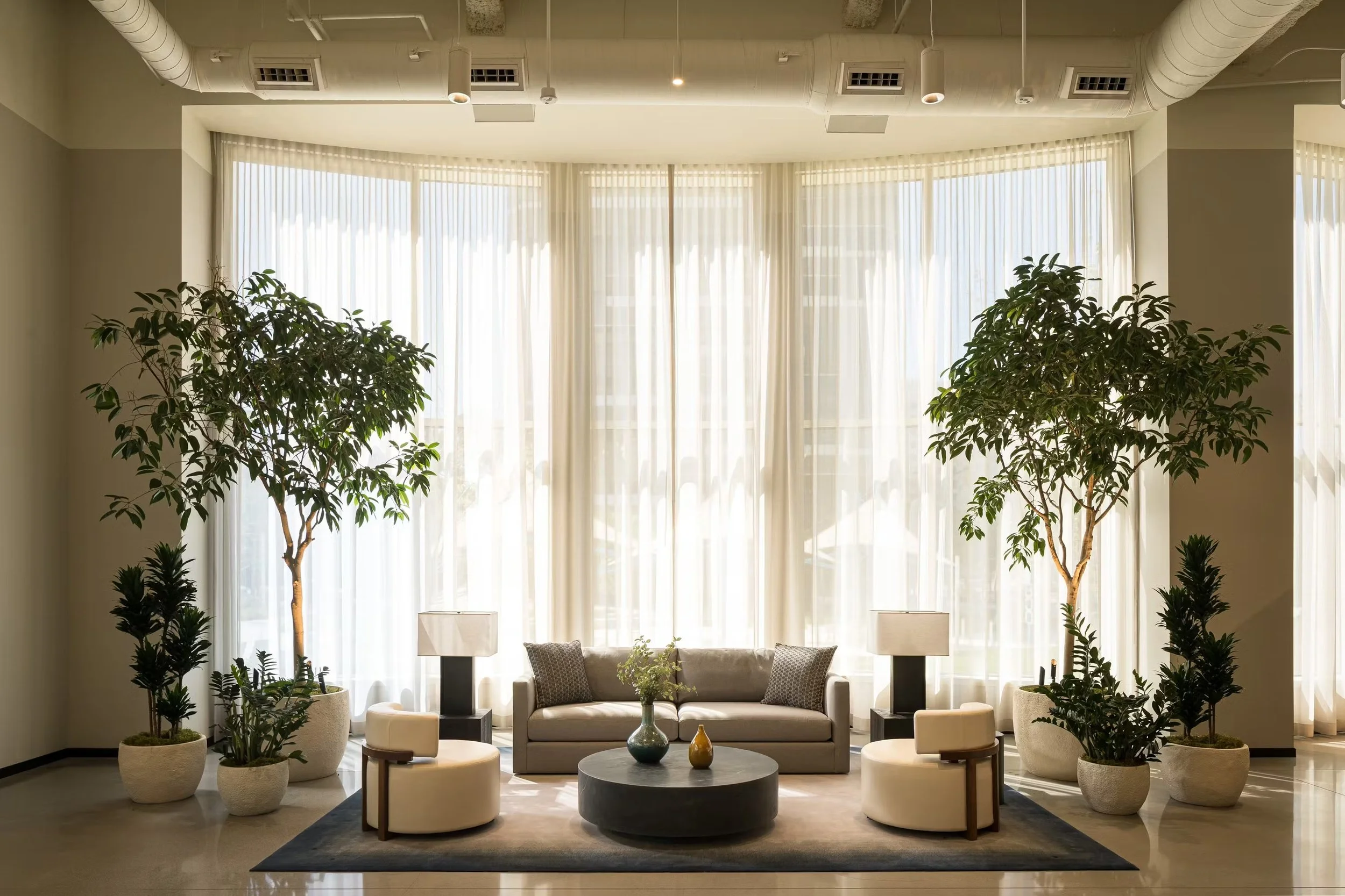 Elegant living room with a large, curved window, beige sofa, two white chairs, and green potted plants, including two tall trees flanking the sofa, with a coffee table in the center, decorated with vases, and table lamps on side tables.