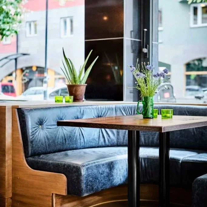 Cozy restaurant corner with a wooden table, a blue velvet booth, a clear glass vase with purple flowers, and a potted aloe plant near a large window showing parked cars outside.
