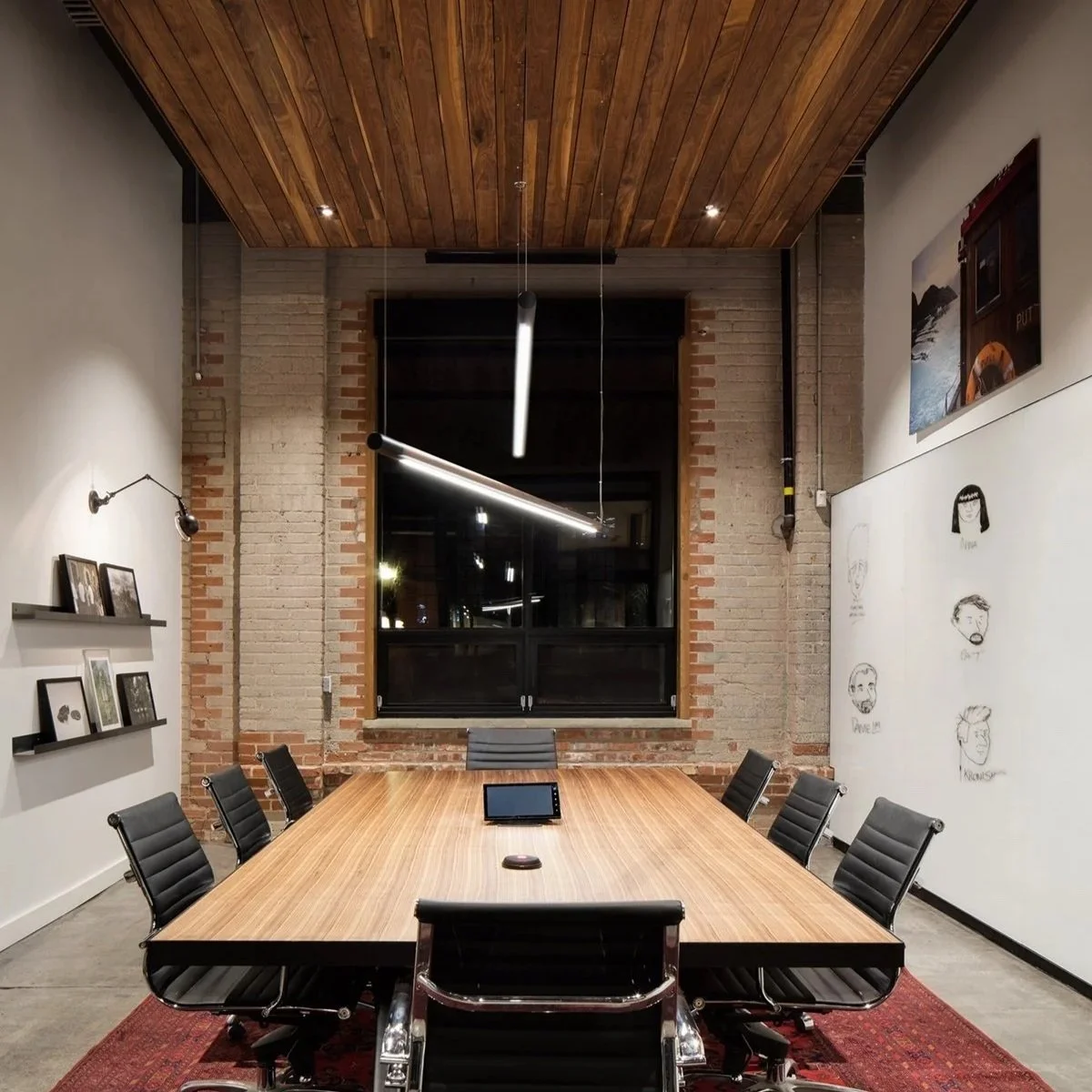 Conference room with a long wooden table, black leather chairs, a tablet and a computer mouse on the table, brick and white walls, hanging lights, large window, artwork on the walls, and a red rug.