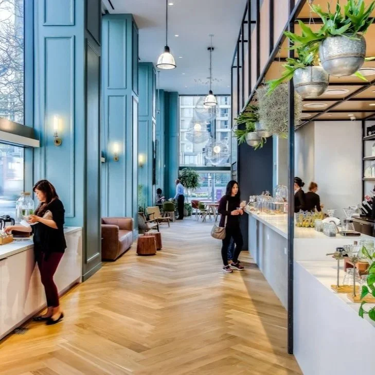Modern cafe interior with large blue walls, wooden floor, hanging pendant lights, and greenery, with people ordering in a coffee shop area.