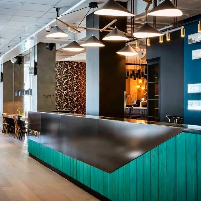 Modern restaurant reception area with a black and teal front desk, hanging black pendant lights, wooden chairs along the left wall, and a patterned wall in the background.