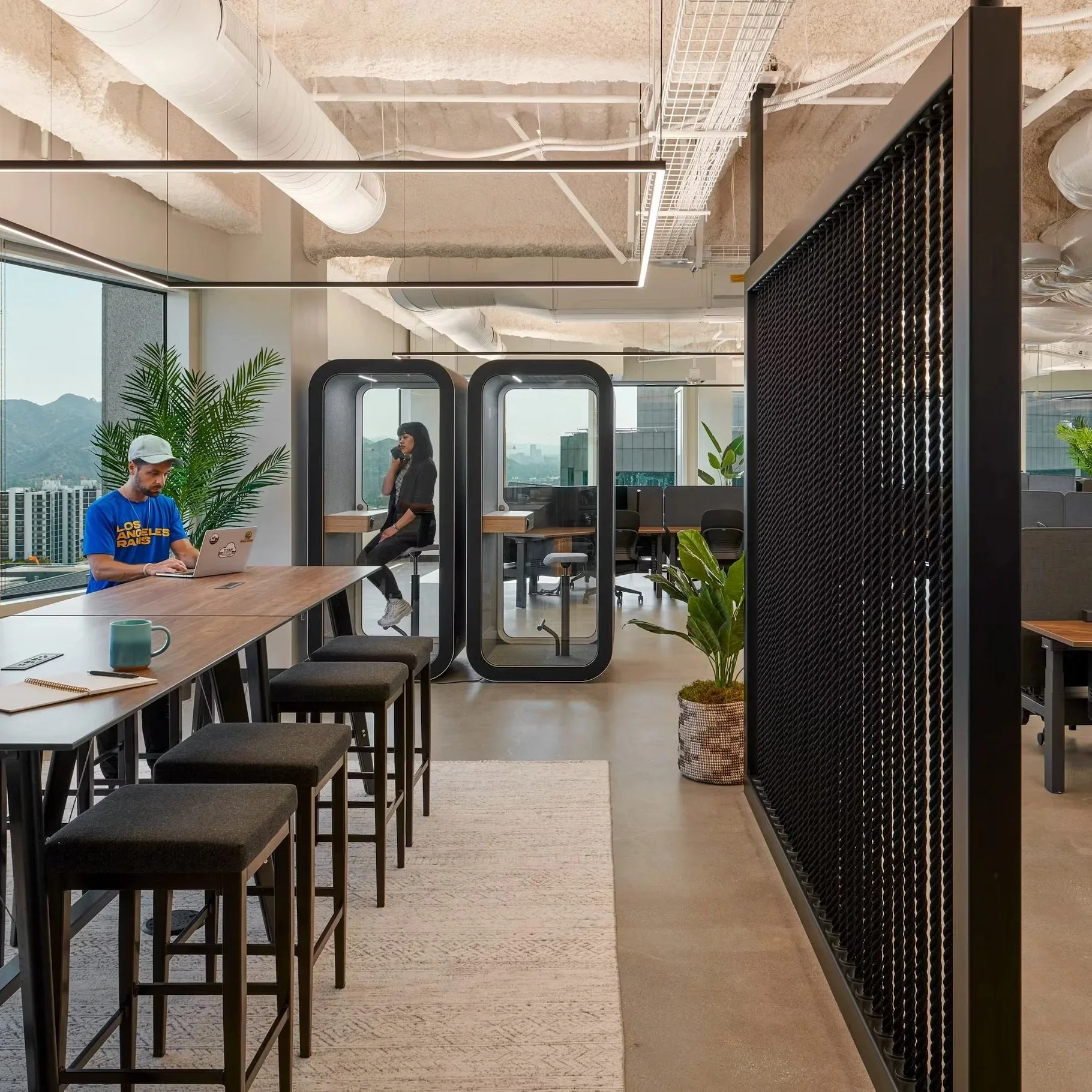 Modern office space with a high table, stools, potted plants, glass phone booths, and workstations in the background, with mountain views through large windows.