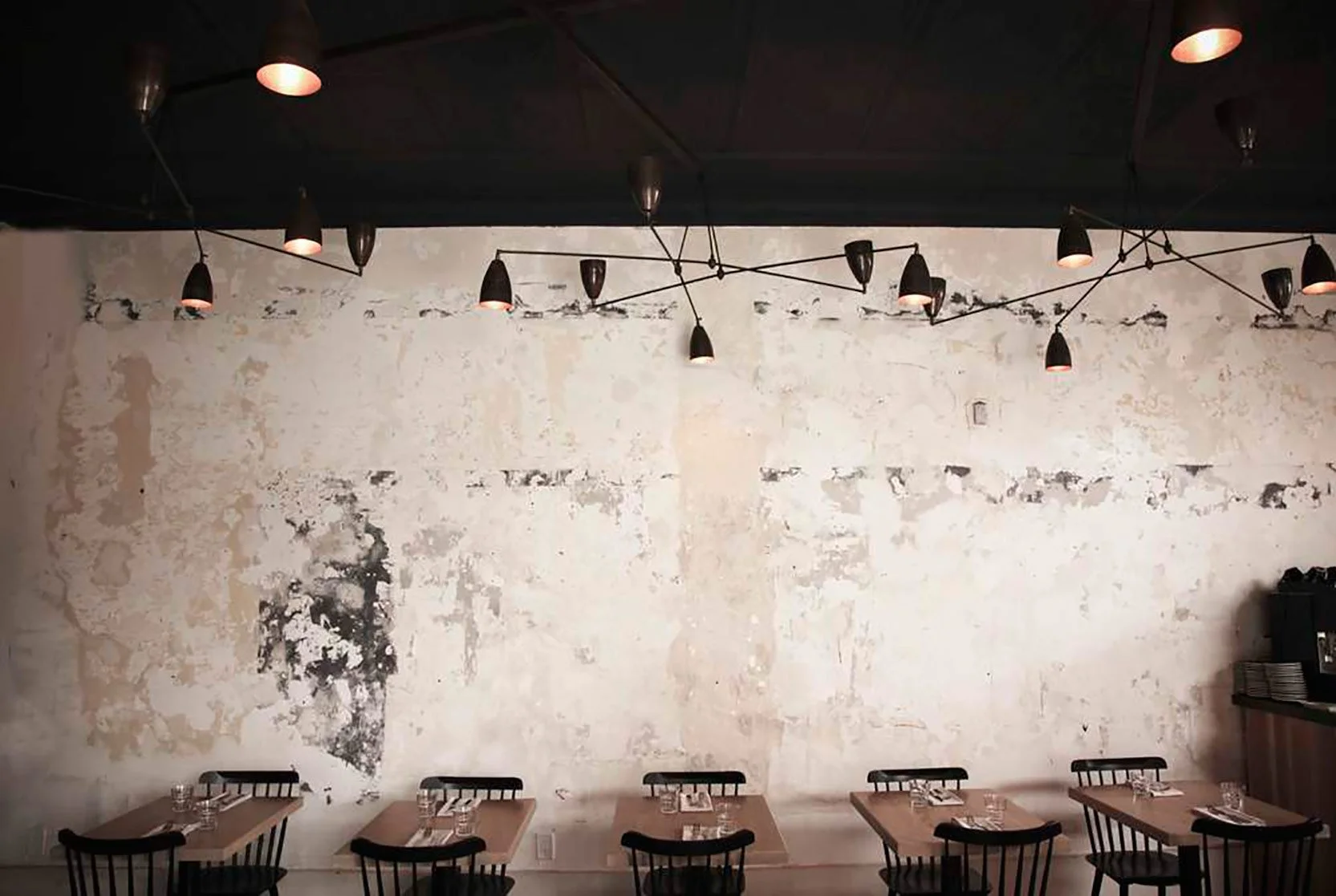 Interior of a minimalist restaurant with a textured off-white wall, black chairs, wooden tables set with glasses and silverware, and modern black pendant lights hanging from the dark ceiling.