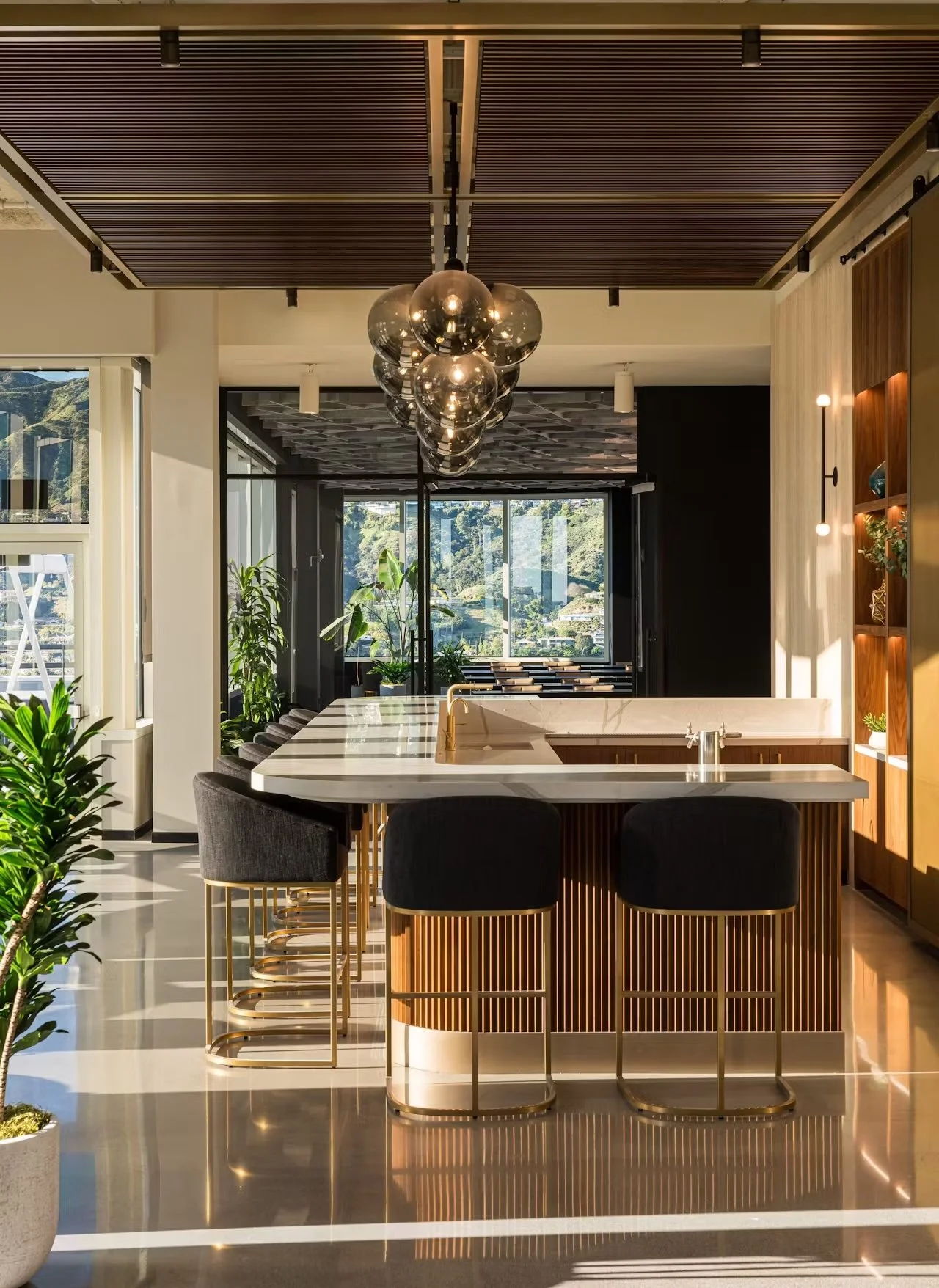 Modern kitchen with a marble island, black stools, green plants, and large windows overlooking a scenic landscape.