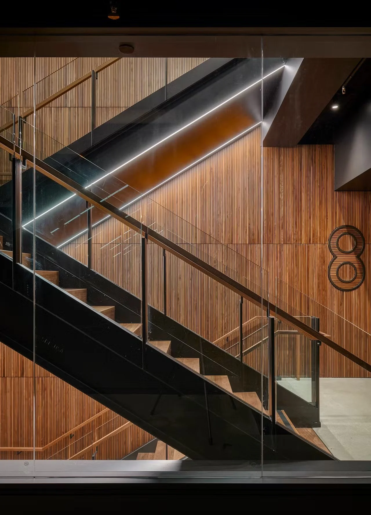 Interior view of a modern staircase with black steps, glass walls, wooden panel walls, and sleek lighting, including the number 80 on the wall.