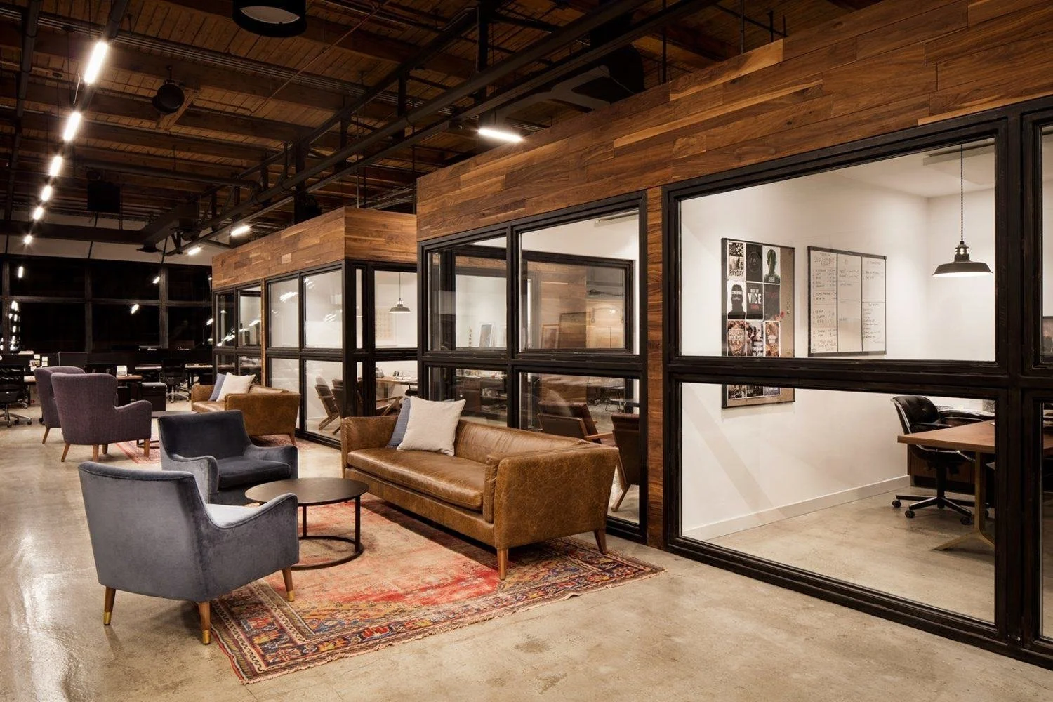 Modern coworking space with glass-walled offices, leather and upholstered chairs, a colorful area rug, and wooden accents on the walls and ceiling.