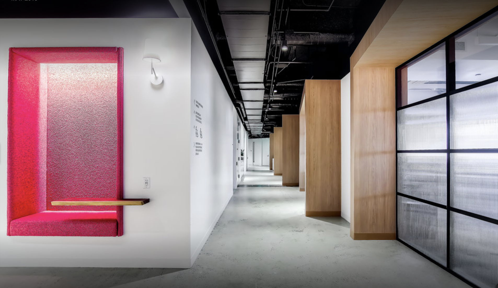 Modern office corridor with a bright pink booth, white walls, wooden panels, and black ceiling with exposed ductwork.