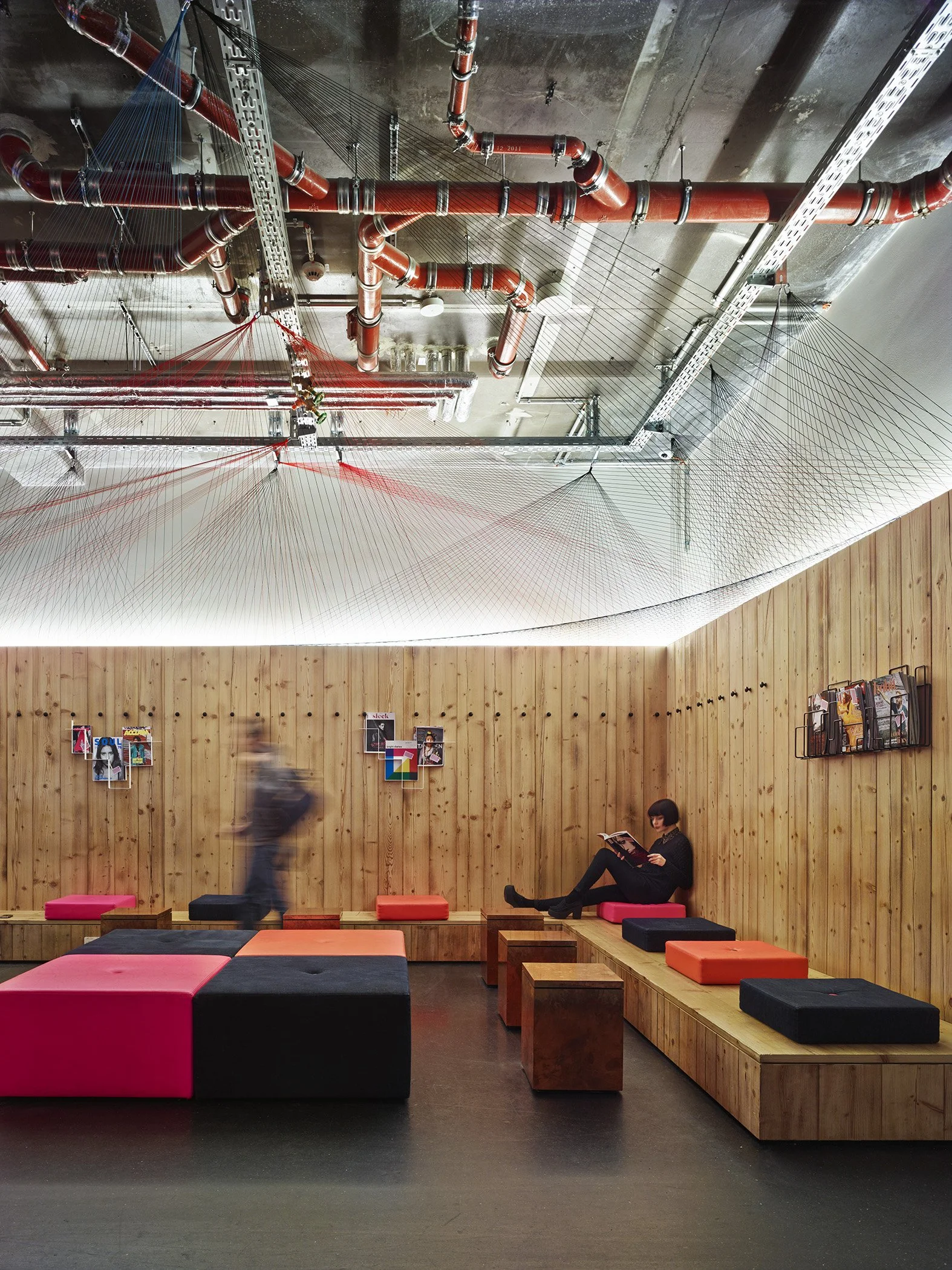 A modern lounge with wooden walls and colorful cushioned seating. A woman is sitting and reading a magazine, while a person walks by blurred due to motion. The ceiling features exposed pipes and a metal grid structure.