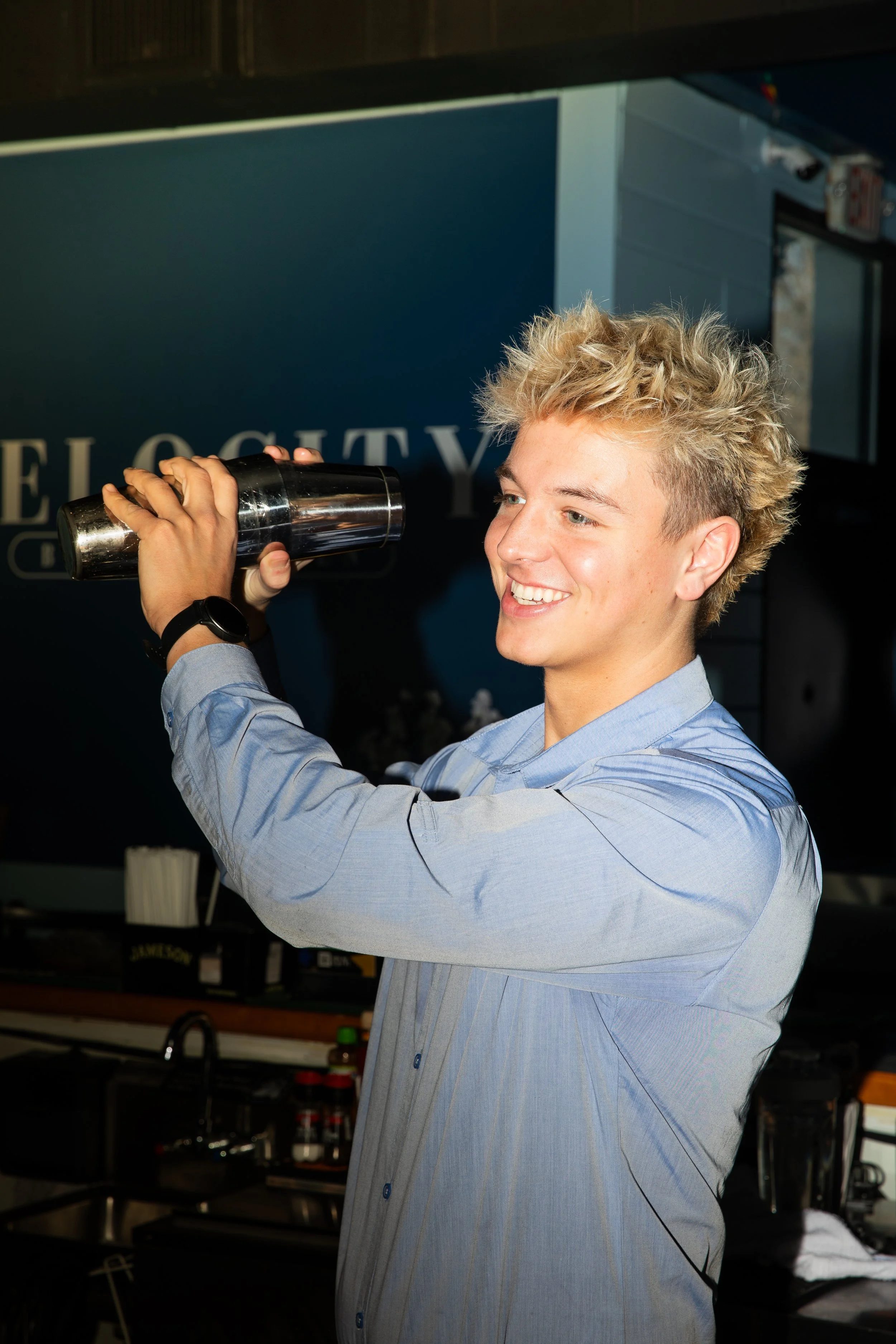 Bartender shaking a metal cocktail shaker behind a bar, with glassware and bottles visible in a dimly lit restaurant setting.