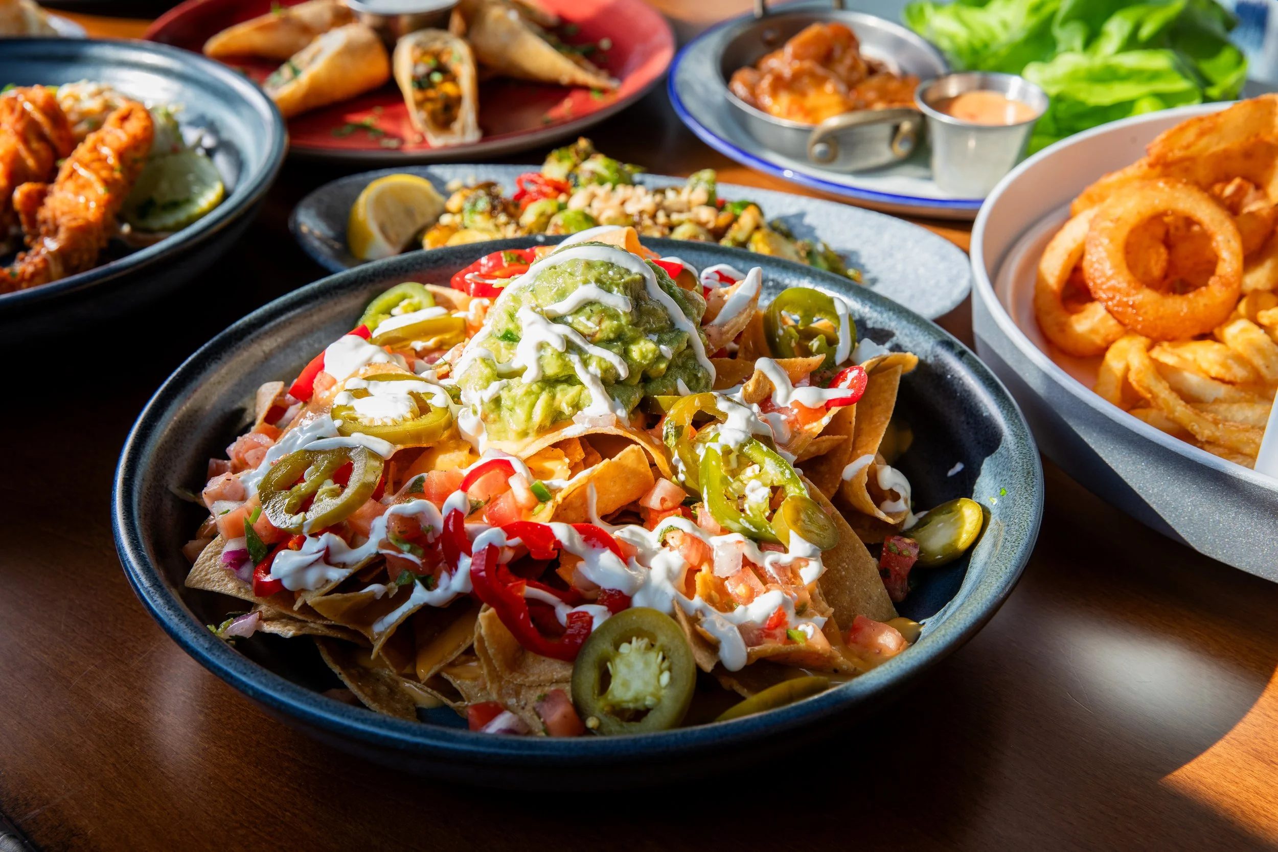 Loaded nachos topped with cheese, jalapeños, tomatoes, sour cream, and guacamole, served with other appetizers and sides on a restaurant table.
