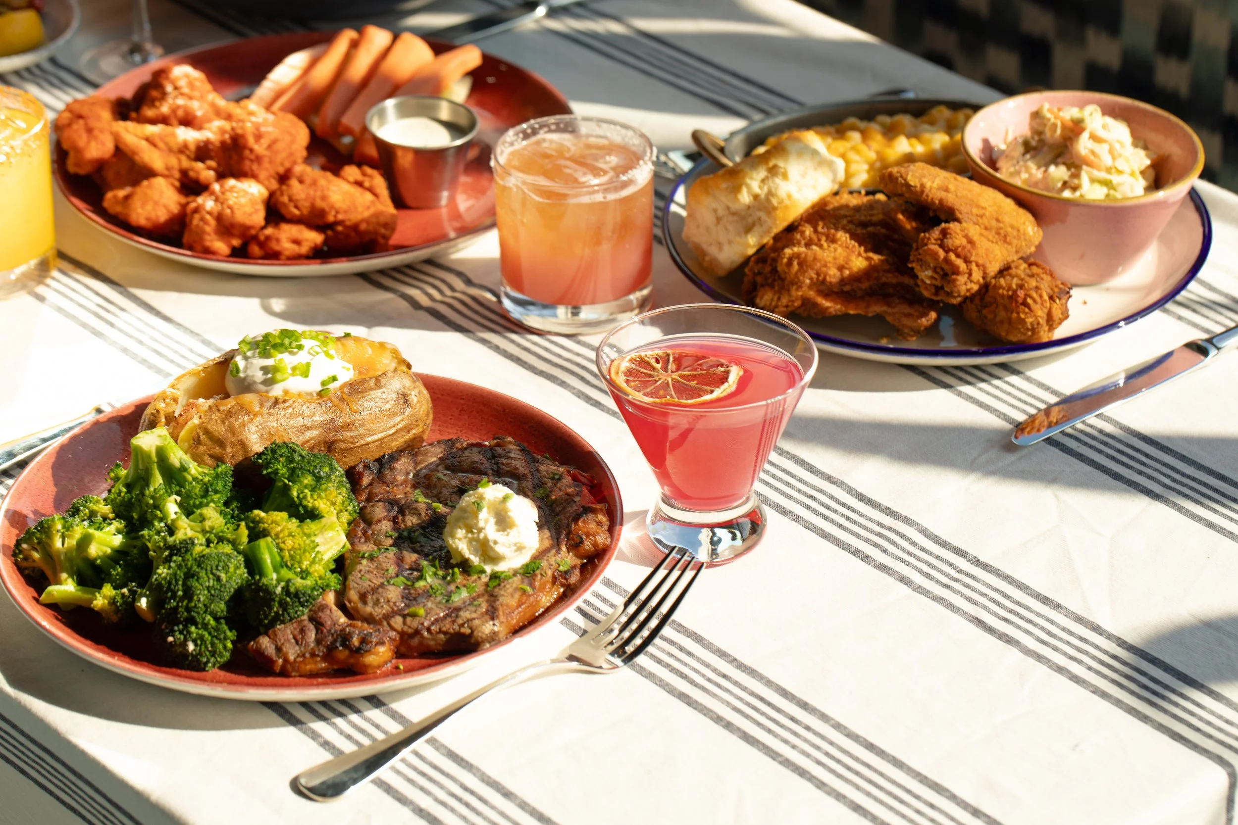 Table spread featuring steak with broccoli and baked potato, fried chicken with sides, cocktails, and wine set for a shared dining experience.