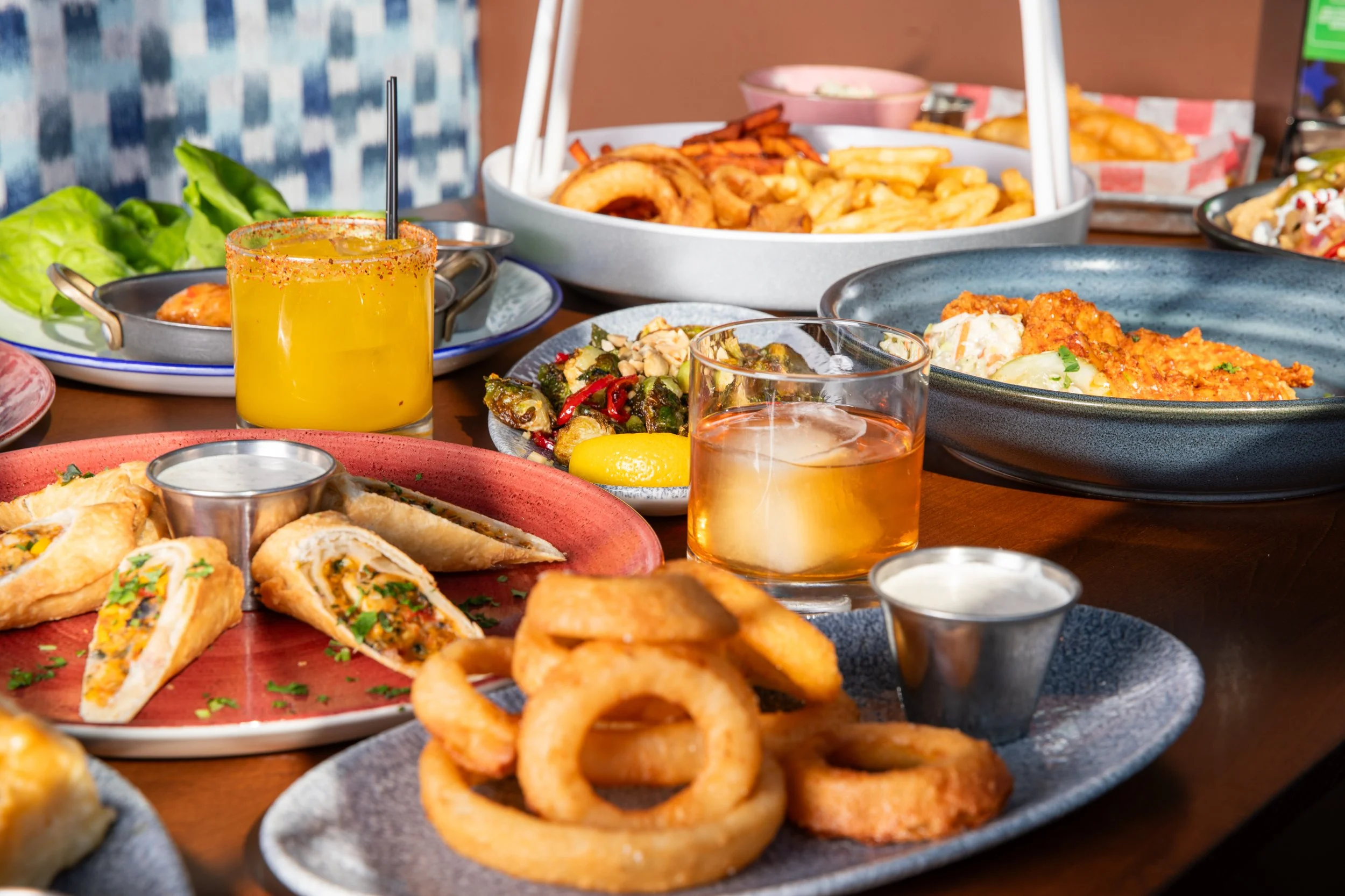 Restaurant table covered with appetizers, onion rings, wraps, sauces, and cocktails, with plated meals visible in the background.