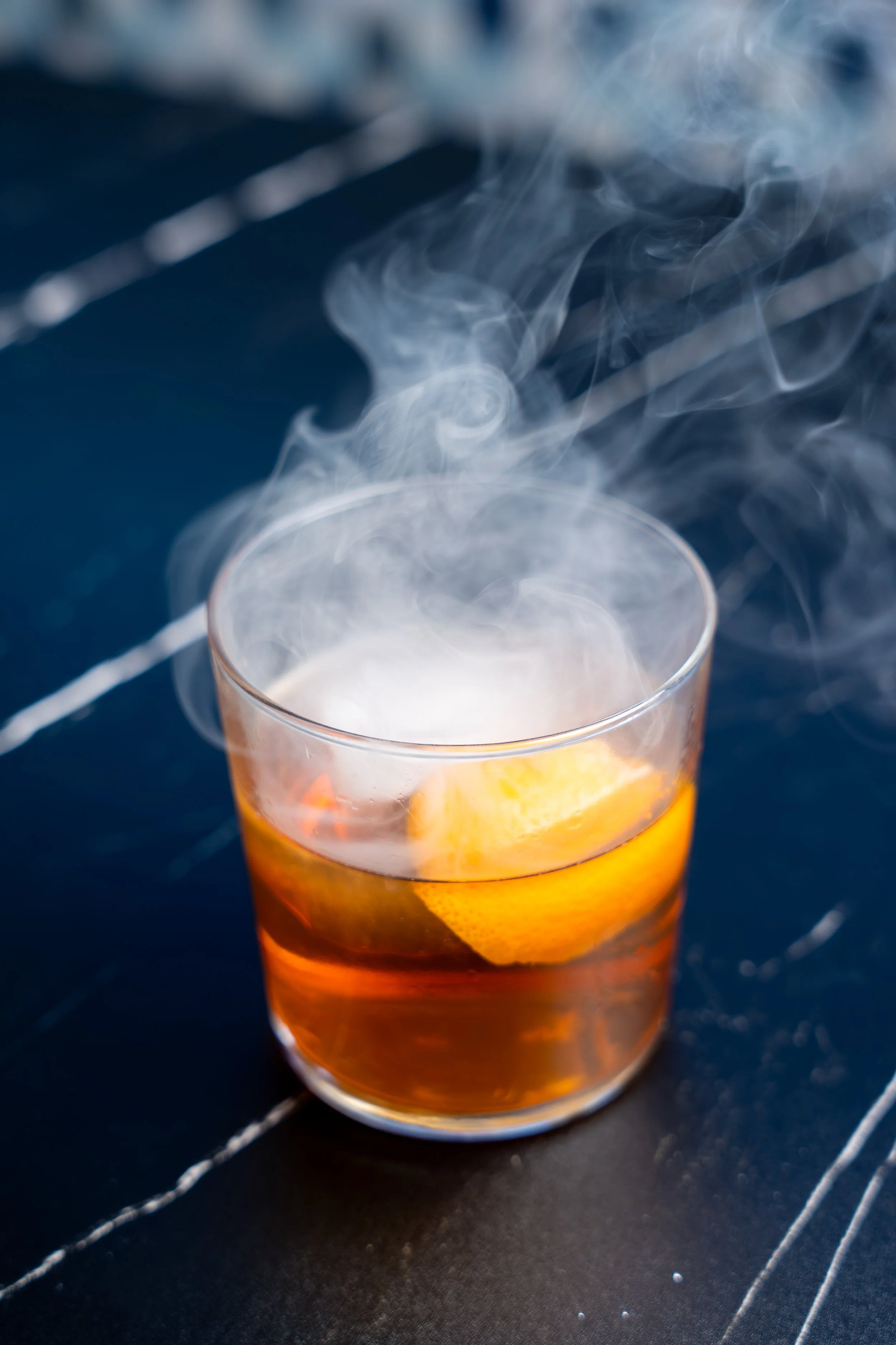 Glass of amber-colored cocktail with citrus peel, surrounded by swirling smoke.
