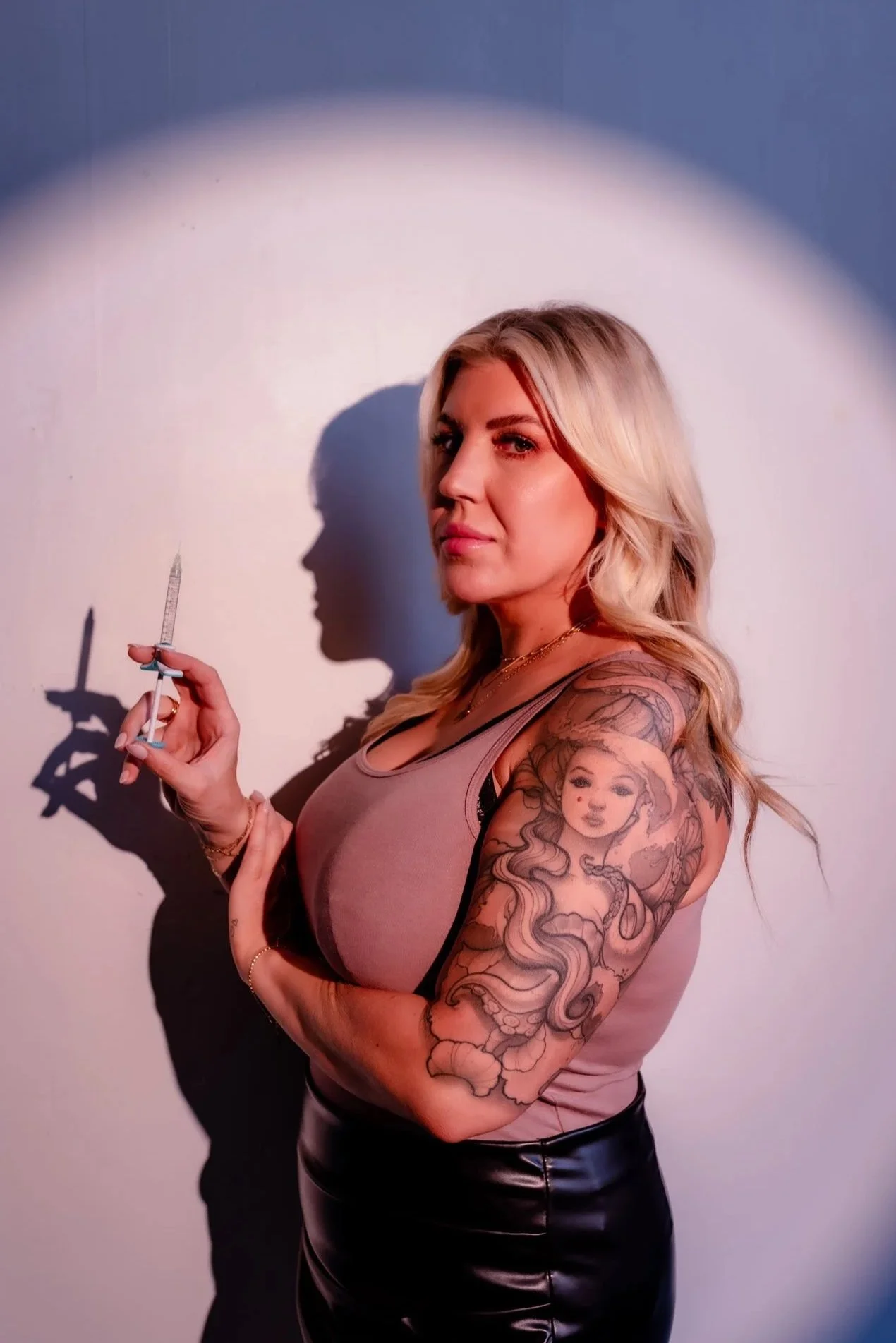 A woman with blonde hair and tattoos on her arm, holding a syringe, standing against a wall with a spotlight casting her shadow on the wall.