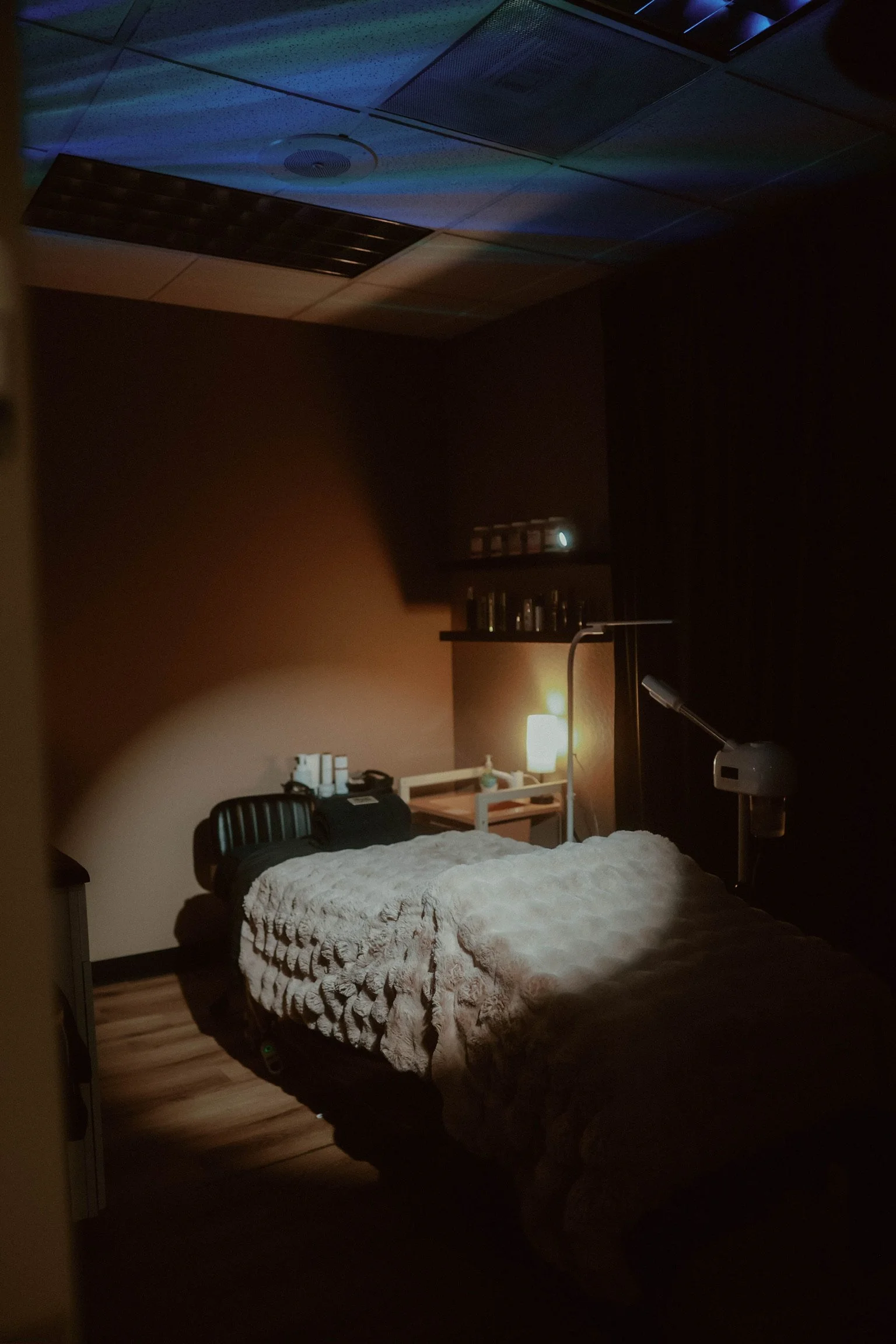 A dimly lit bedroom with a bed covered by a textured, light-colored blanket, next to a small nightstand with a lamp. There is a chair, several bottles, and electronic devices near the bed. The ceiling has acoustic tiles and ventilation panels, with some colored light reflections.