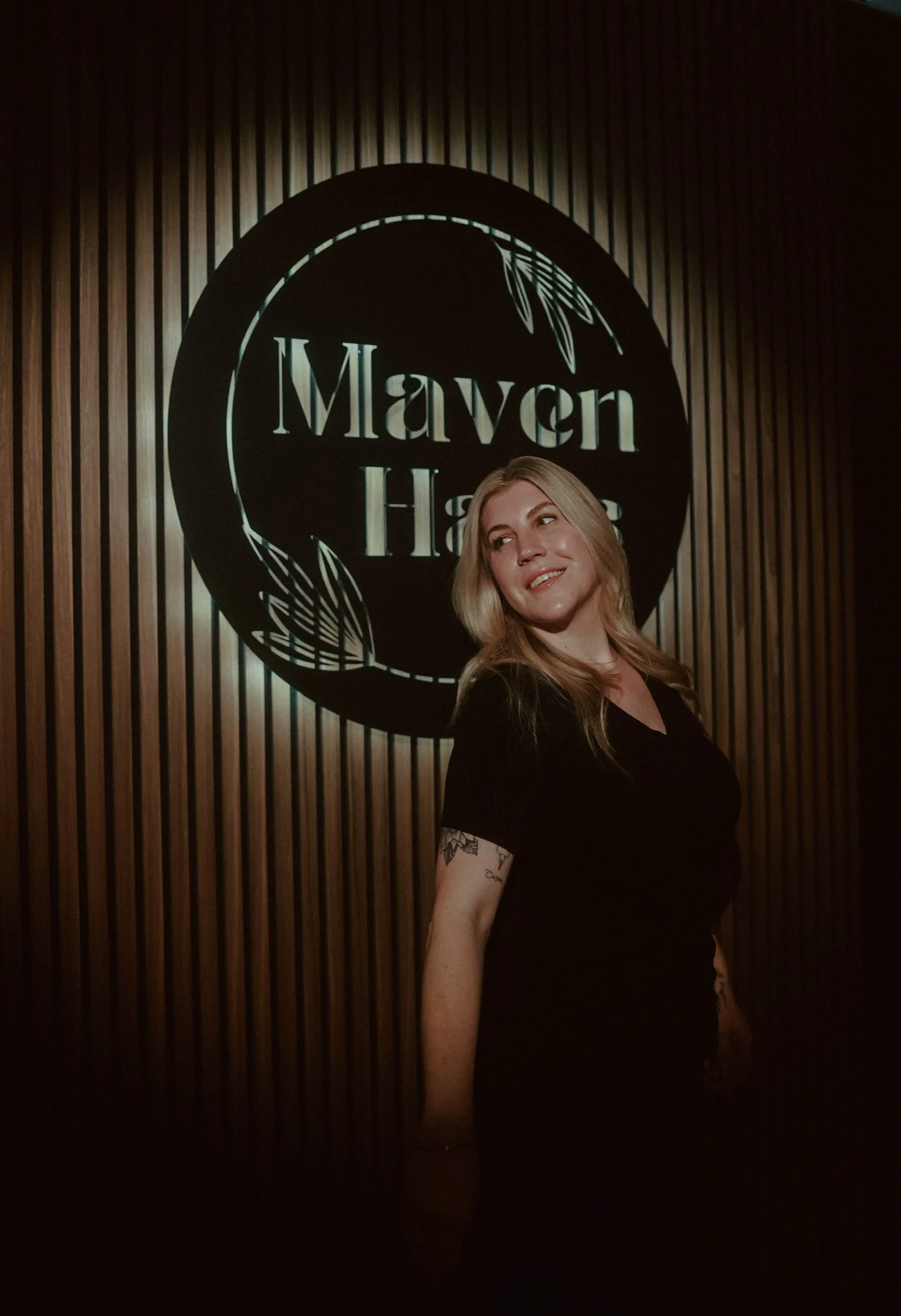 Ashley Walkup BSN RN aesthetic nurse injector and founder of Maven Haus Vacaville standing in front of logo wall.