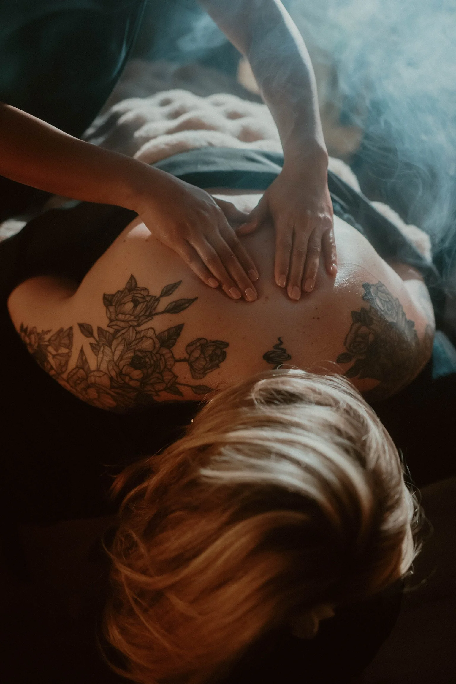 A person receiving a massage, with hands on their back, tattoos on their shoulder and blonde hair displayed.