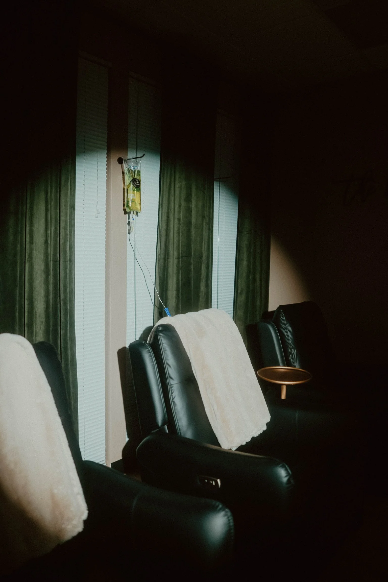 A row of black leather chairs with white towels draped over the backrests in a dimly lit room, with three windows with closed blinds and green curtains in the background, one window has an IV drip hanging nearby.