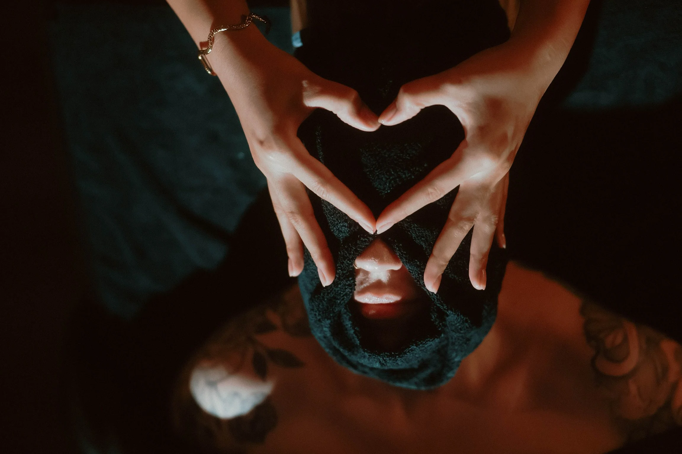 Person's hands forming a heart shape over their face, with dark background.