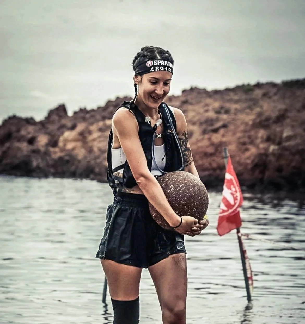 Une femme sportive en tenue de course, portant une bandeau noire, sourit tout en tenant une boule d'entraînement, sur un fond de lac et de rochers, avec un drapeau rouge derrière elle.