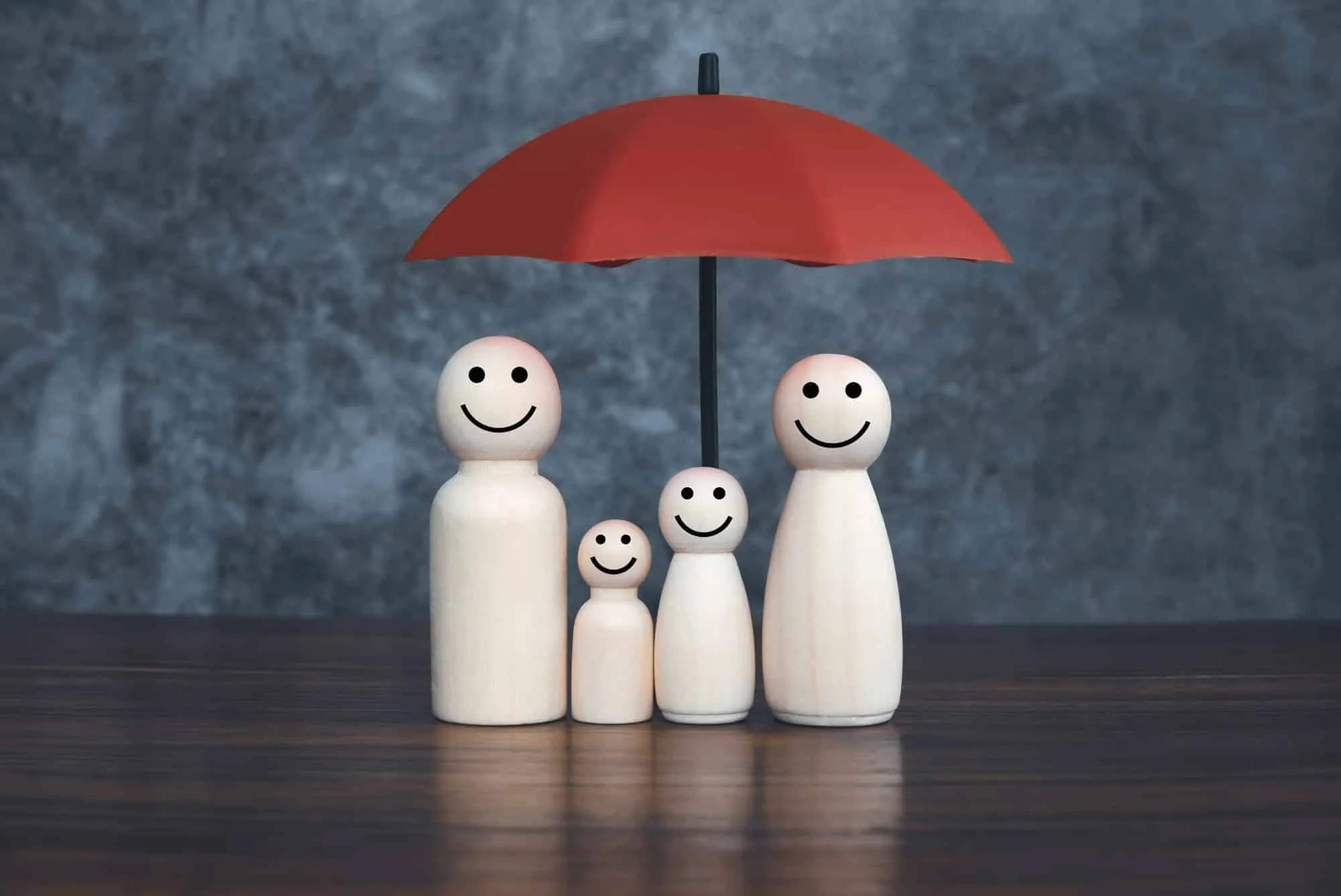 Four wooden figurines with smiley faces under a red umbrella on a wooden surface against a gray textured background.