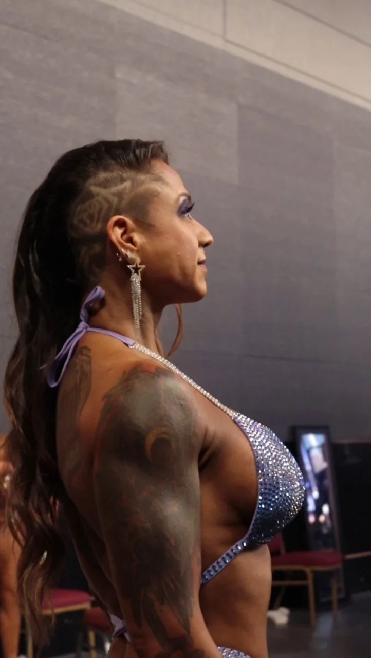 Side profile of a woman with dark long hair styled with a shaved side, wearing large star-shaped earrings, a sparkling rhinestone top, and purple straps with a bow. She has visible tattoos on her upper arm.