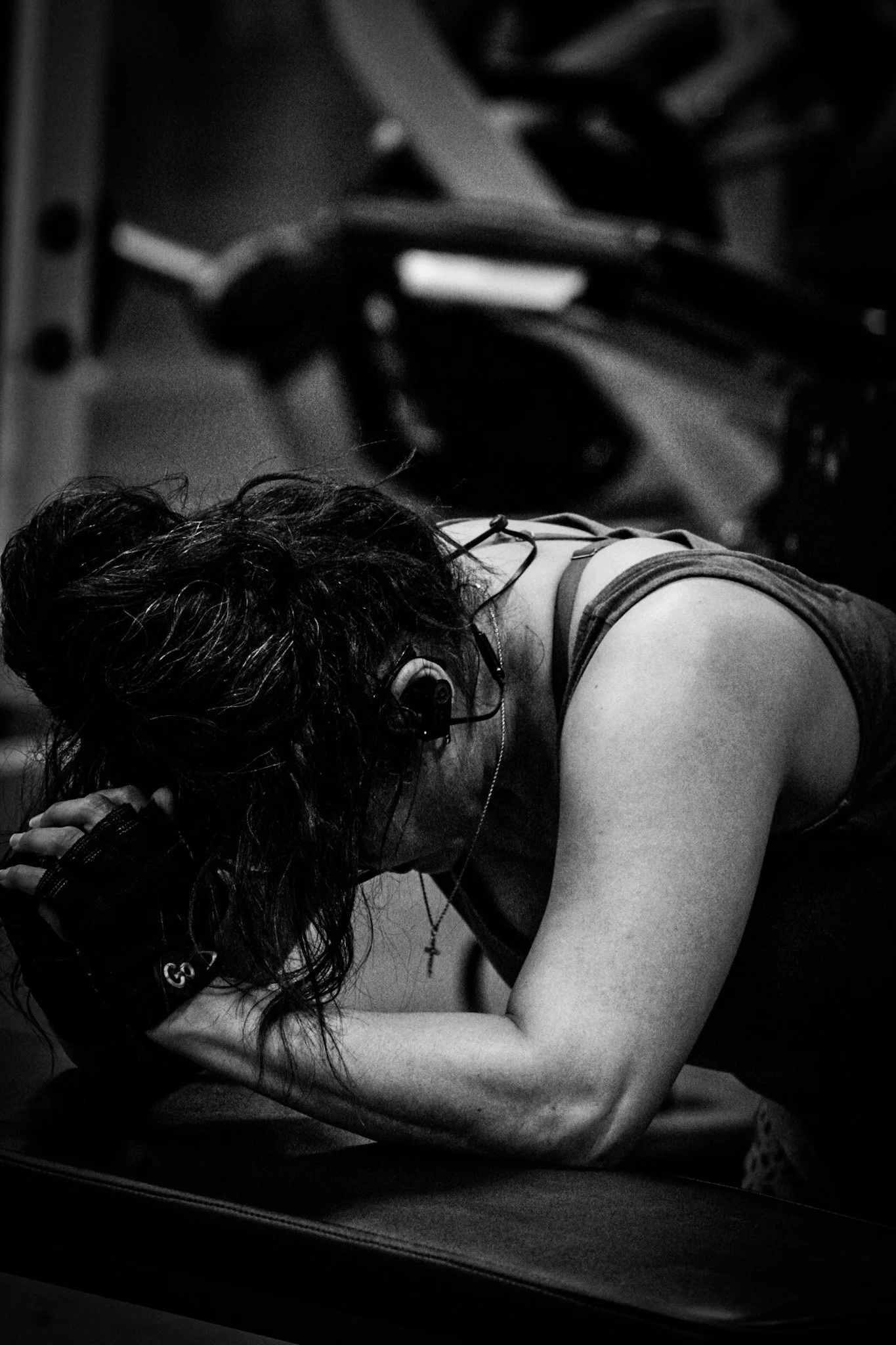 A woman with long hair, wearing a sleeveless top, is resting her head on her folded arms on a bench in a gym, appearing tired or exhausted.